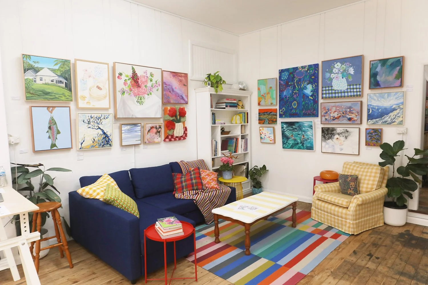 Living room with gallery wall of colorful paintings, navy sofa with patterned pillows, yellow armchair, wooden coffee table on a rainbow-striped rug, and various potted plants.
