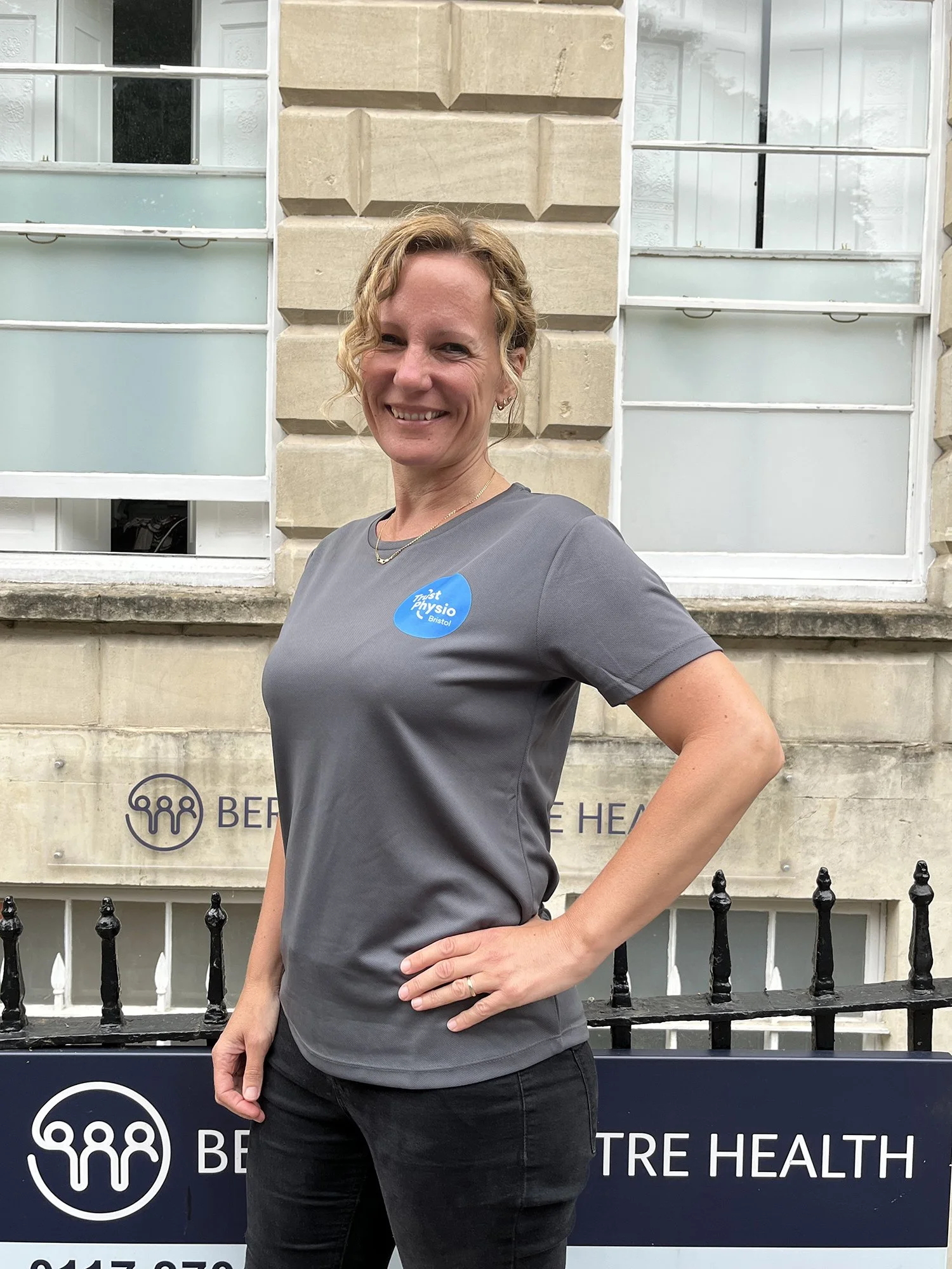 A photo of Claire Lynch - Physiotherapist wearing a gray T-shirt with a blue circular Trust Physio Bristol logo. She has her left hand on her hip and is smiling. The background includes a stone wall and partially visible windows.