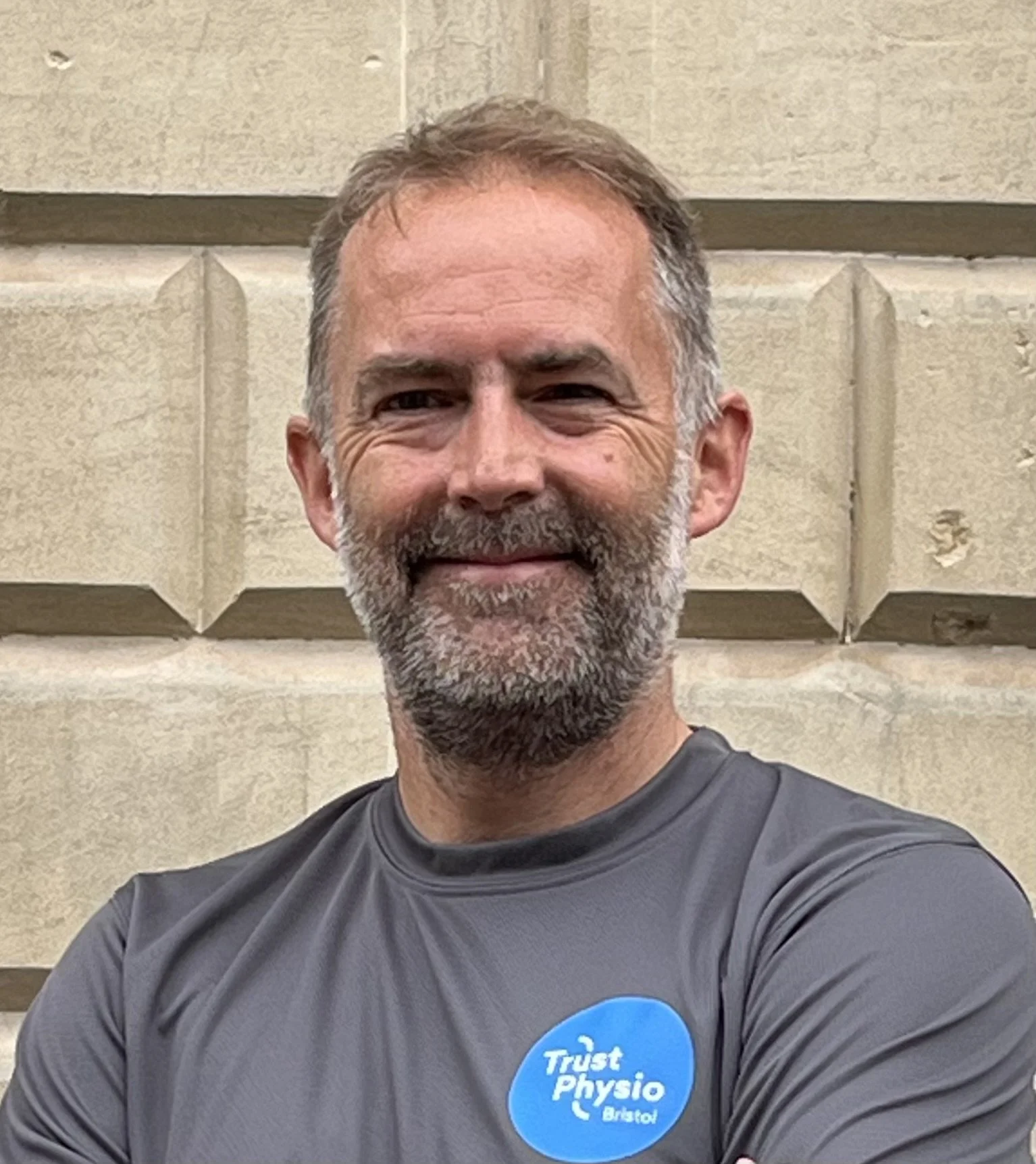 A photo of Owen Lynch - Physiotherapist wearing a gray T-shirt with a blue circular Trust Physio Bristol logo. He is crossing his arms and smiling. The background includes a stone wall and partially visible windows.