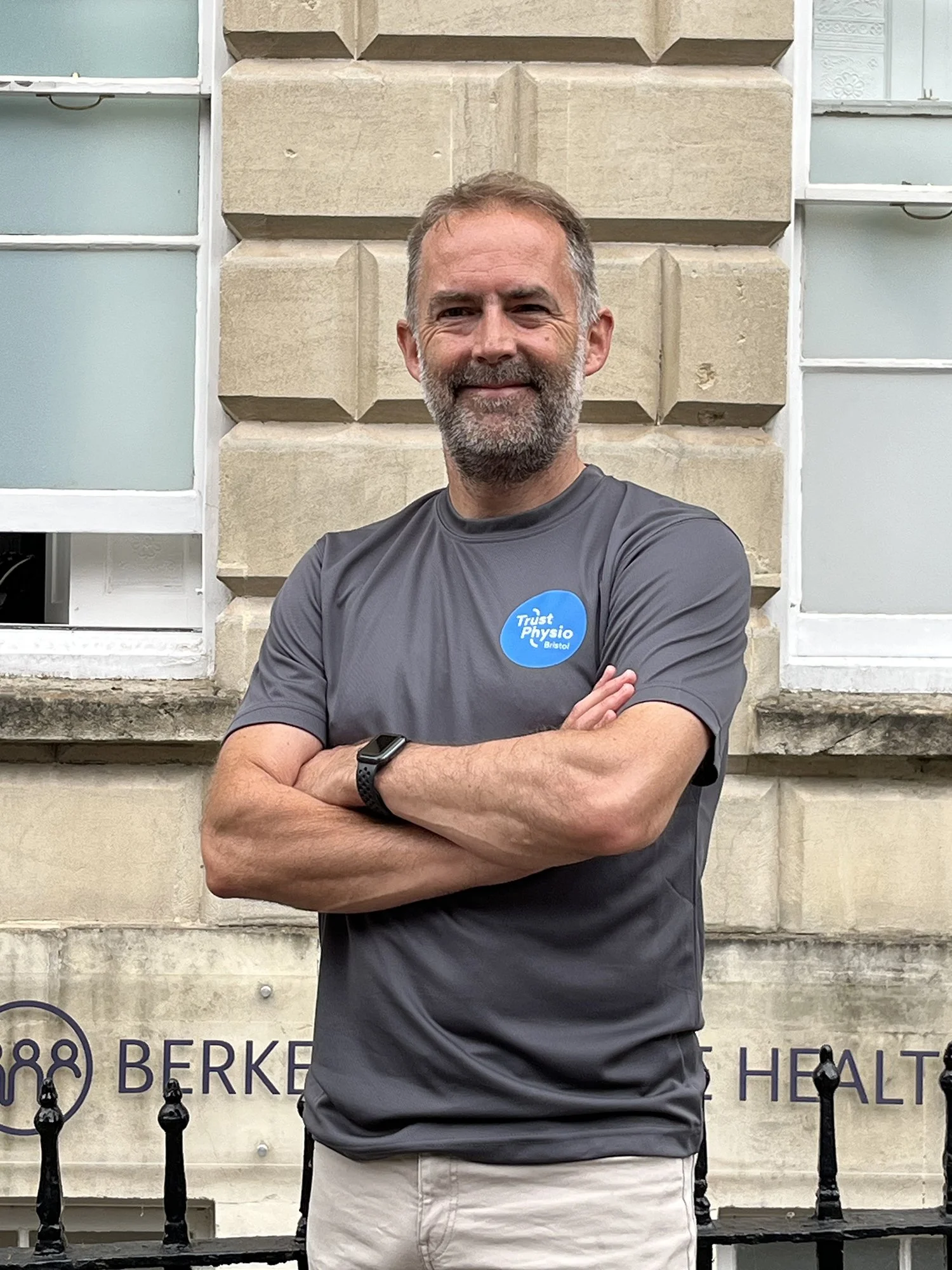 A photo of Owen Lynch - Physiotherapist wearing a gray T-shirt with a blue circular Trust Physio Bristol logo. He is crossing his arms and smiling. The background includes a stone wall and partially visible windows.