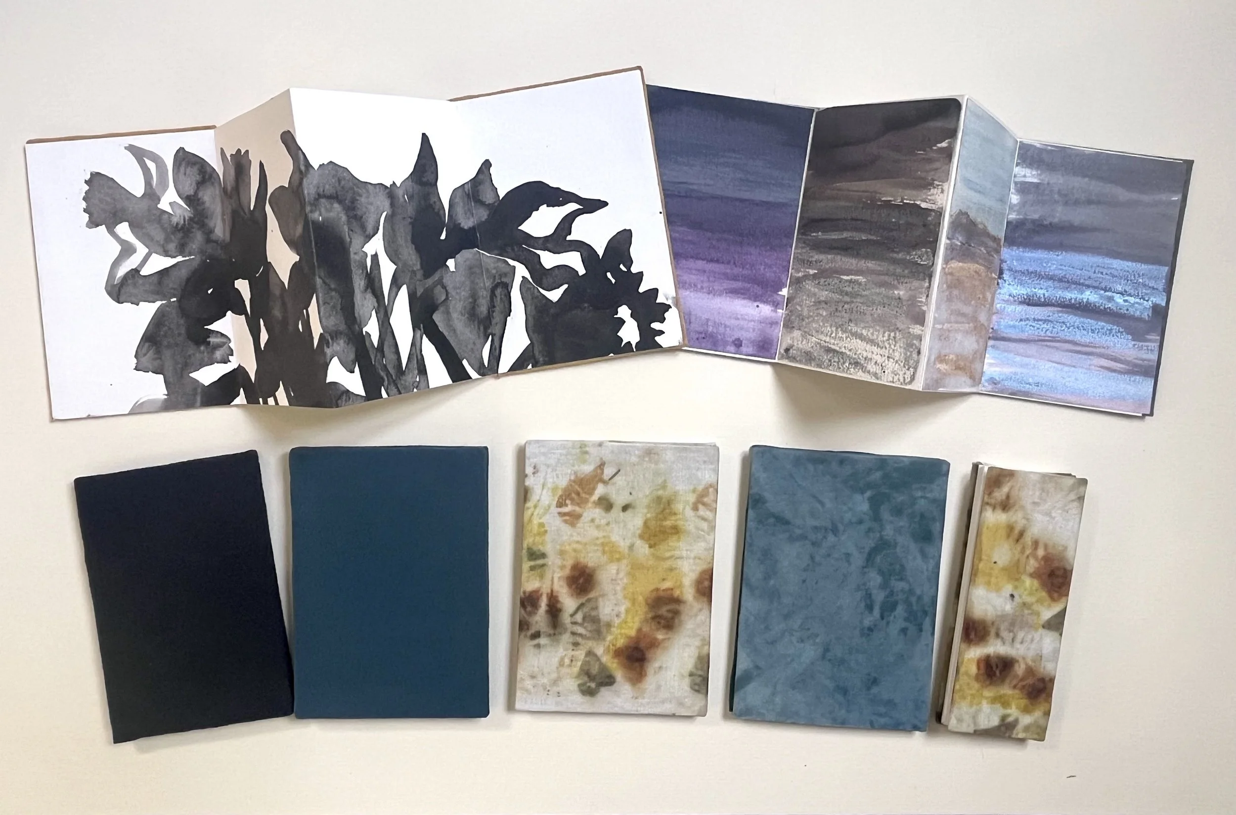 A variety of watercolor art pieces displayed on a wall, including a black and white botanical pattern and abstract landscape scenes in shades of purple, blue, and earth tones.