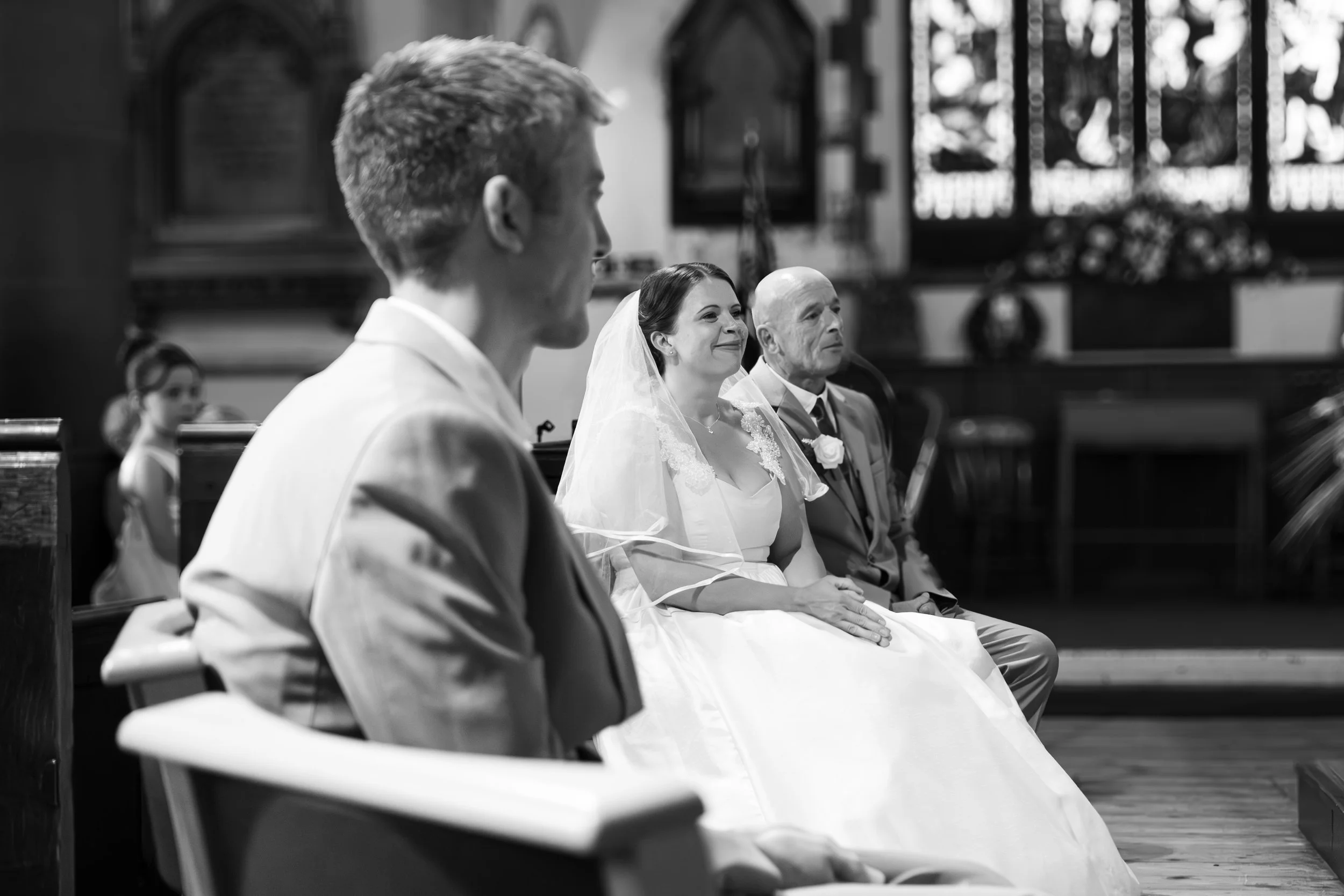 Black and white photo of a wedding ceremony inside a church. The bride is sitting in the middle, smiling, wearing a bridal veil and gown. An older man, likely her father, is sitting to her right. To her left, a groom is seated, turned slightly away f