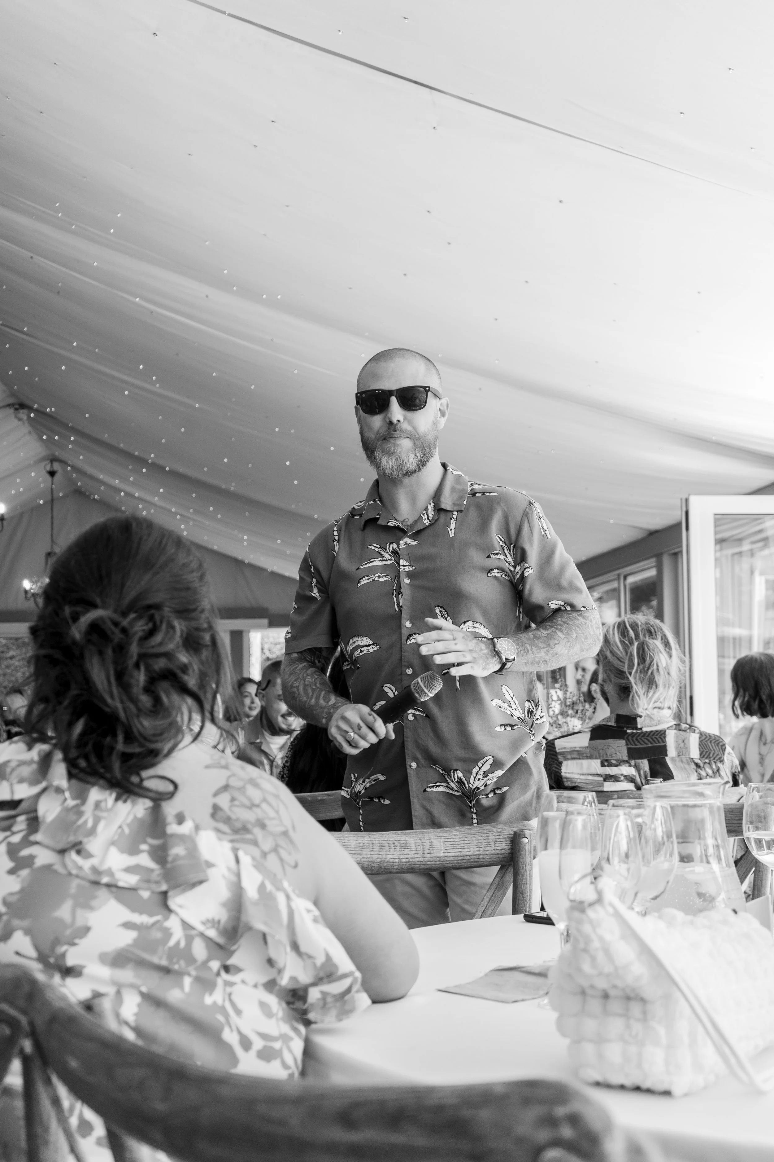 A man with a beard, sunglasses, and tattoos, holding a microphone, stands at a restaurant or event space with people seated at tables, engaged in conversation.