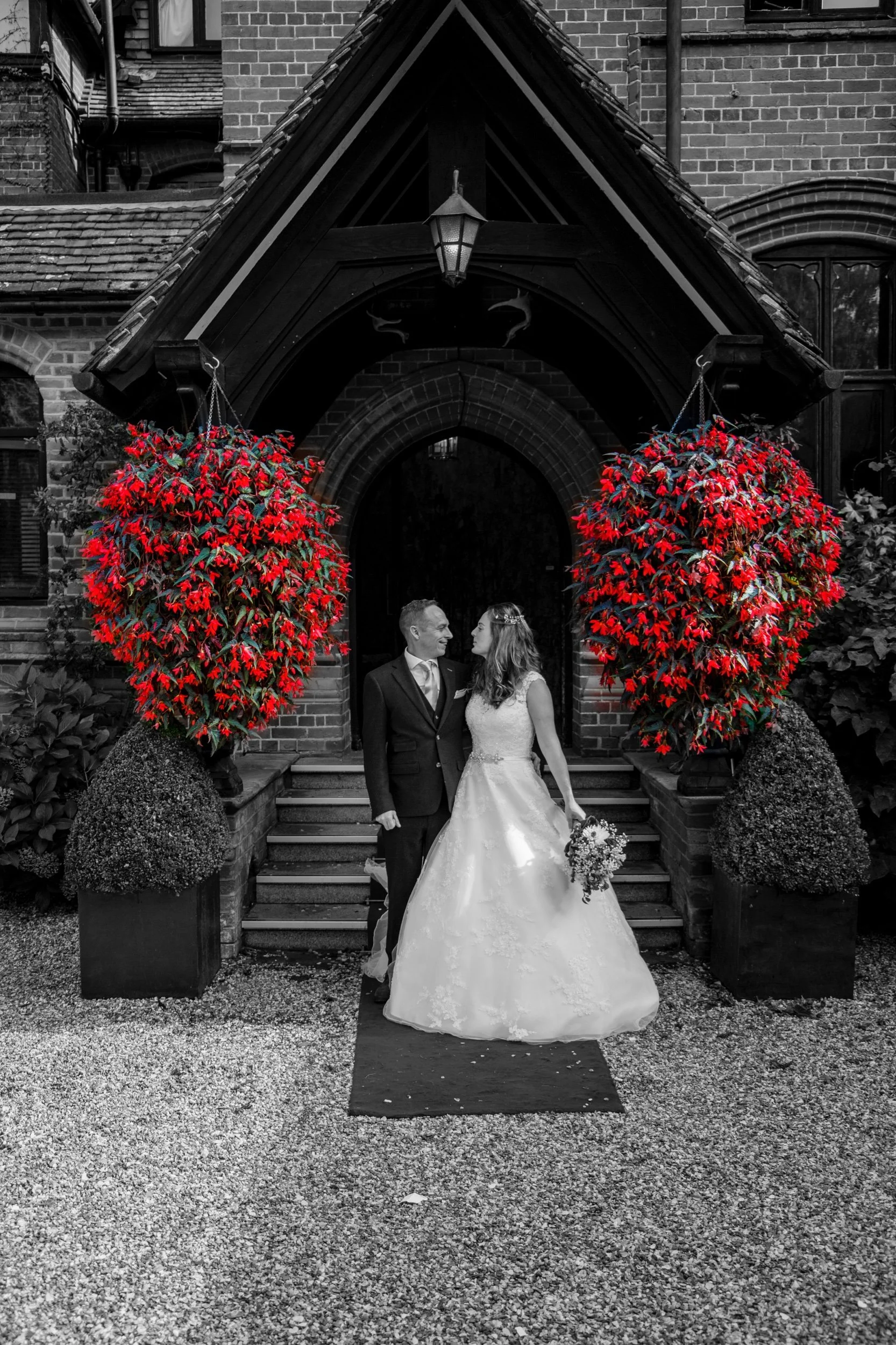 Black and white photograph of a bride and groom standing on steps outside a brick building, surrounded by two large potted plants with red flowers, with selective color highlighting the flowers.