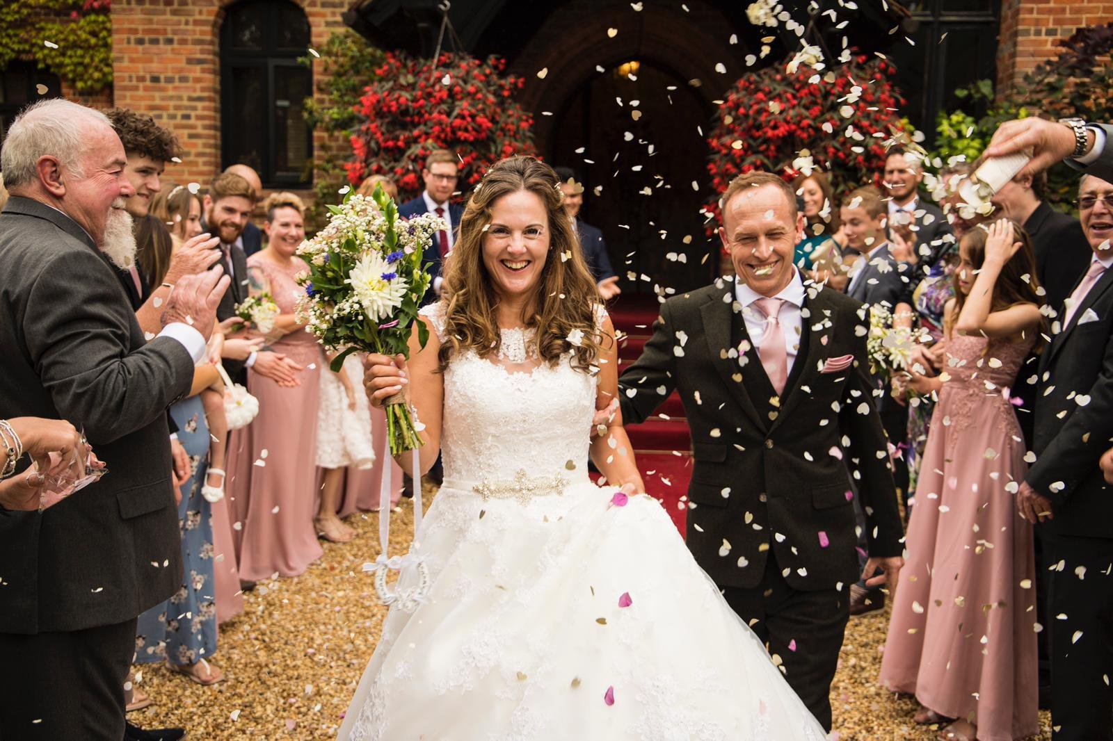 A bride and groom smiling and walking together outside after their wedding, surrounded by friends and family throwing confetti. The bride is holding a bouquet of white and purple flowers, and both are dressed in formal wedding attire. There is a bric