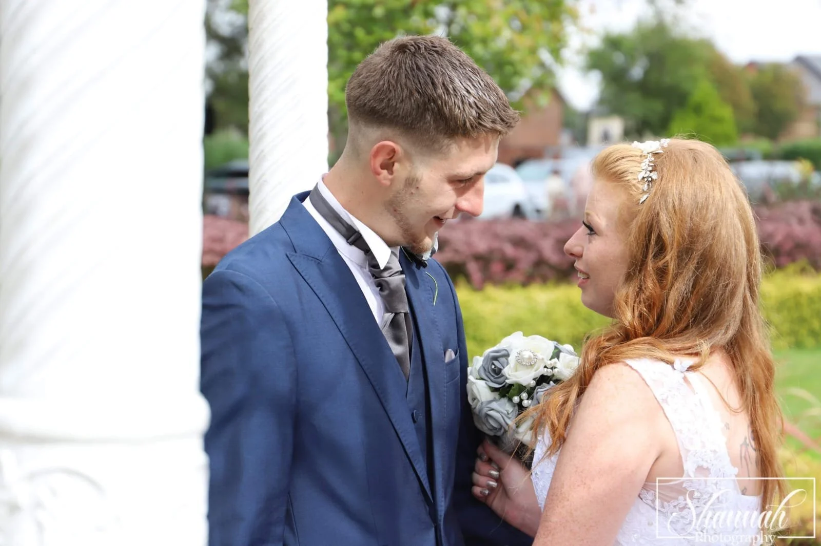 A bride and groom sharing a moment during their wedding ceremony, standing outdoors with a floral bouquet, greenery, and parked cars in the background.