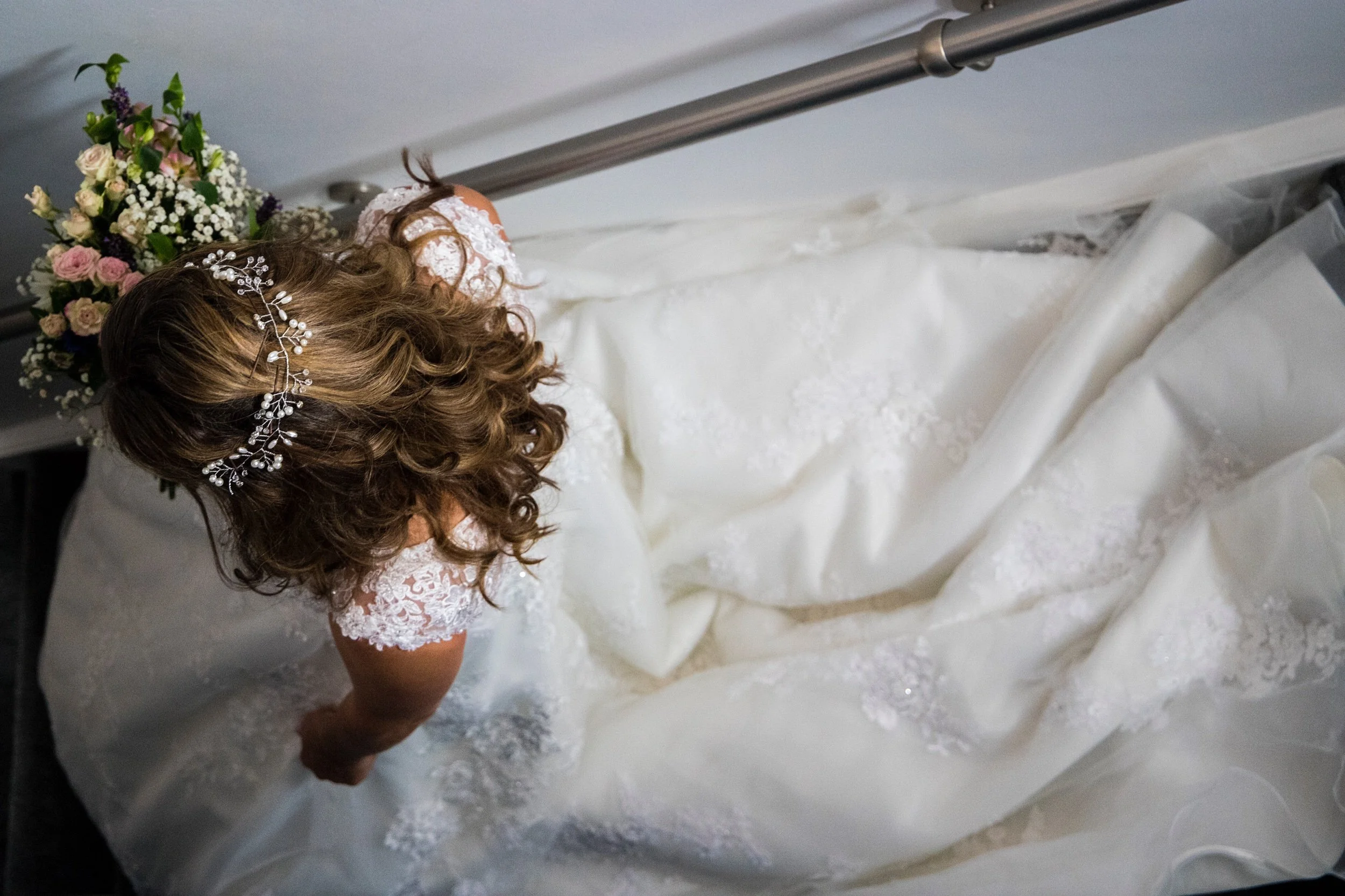 Top-down view of a bride in a white wedding dress with lace details, lying on her side on a bed with a bouquet of flowers nearby.