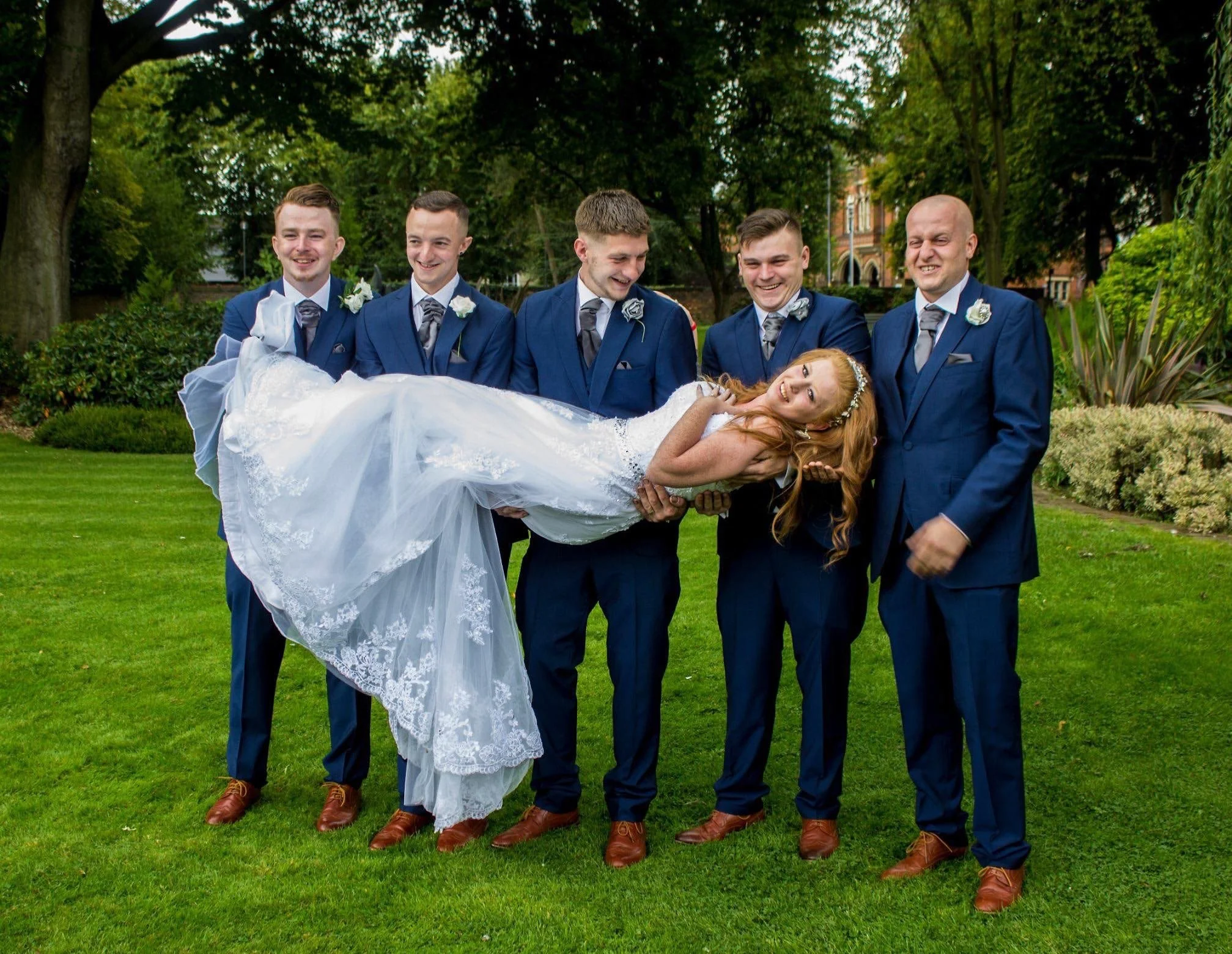 A bride with long red hair wearing a white wedding dress is being held horizontally in the air by six men in blue suits at a park. The men are smiling and standing on a green lawn with trees and bushes in the background.
