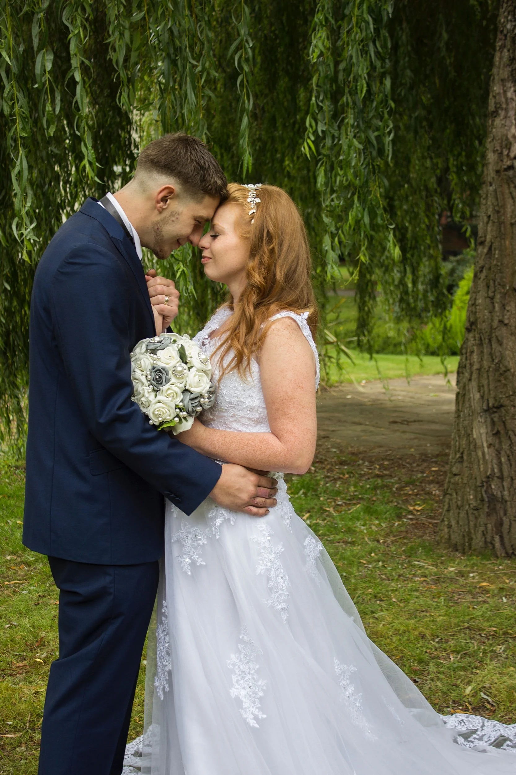 A bride and groom standing close together outdoors, touching foreheads, with green foliage and tree in the background. The bride wears a white lace wedding gown and has long red hair, while the groom is in a dark suit and holds a white bouquet.