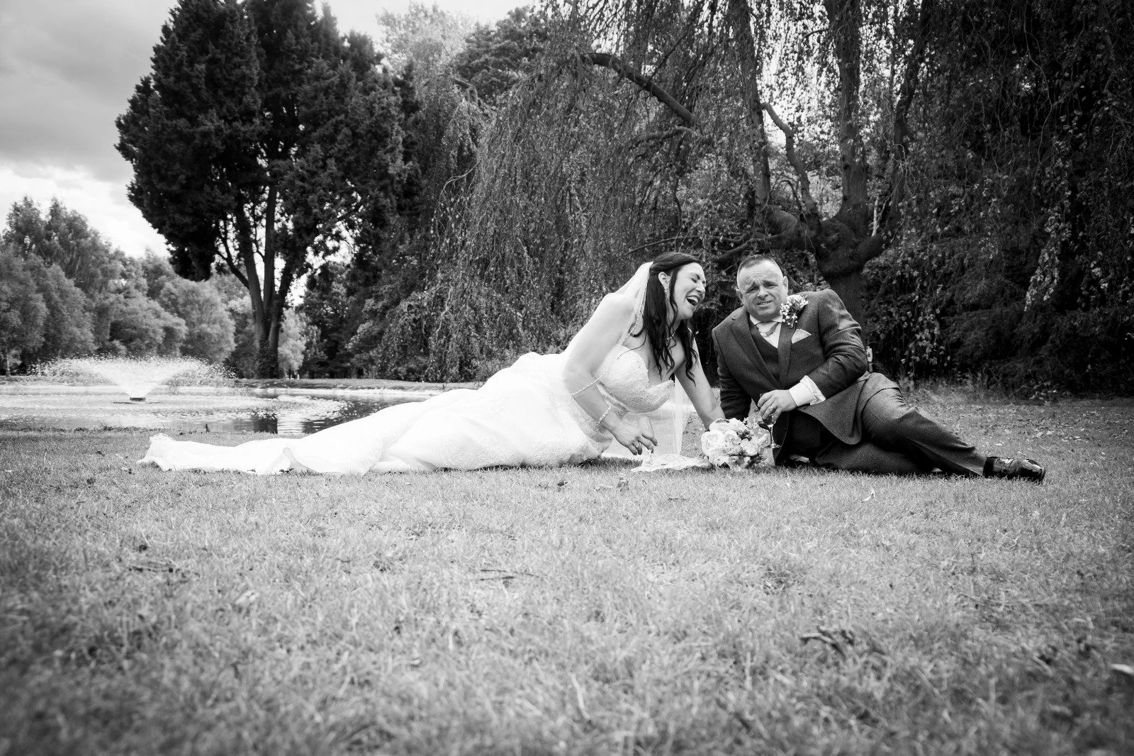 Black and white photo of a bride and groom lying on grass near a pond, laughing and enjoying themselves, with trees and a cloudy sky in the background.