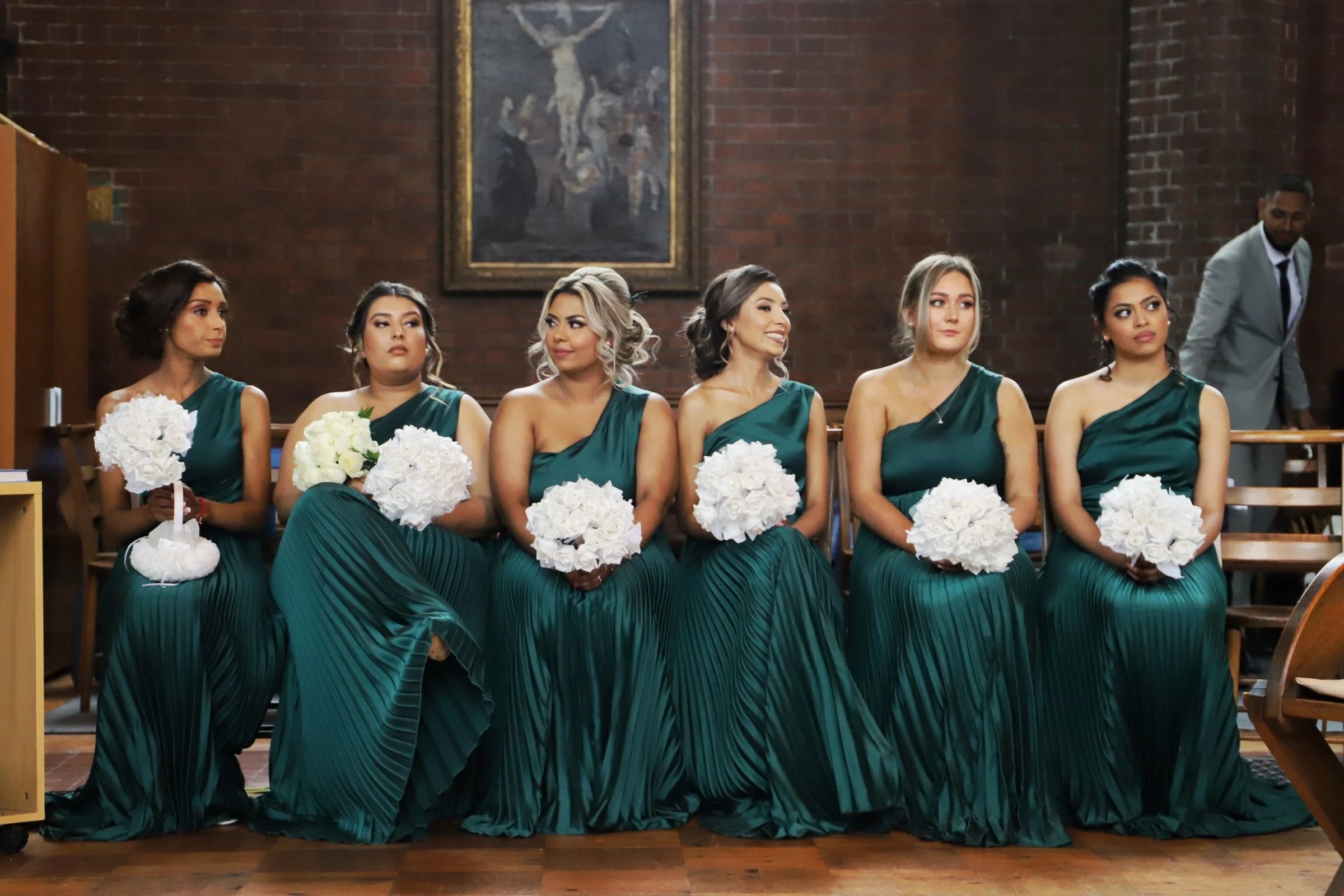 Six bridesmaids sitting in church, wearing matching teal dresses, holding white bouquets, during a wedding ceremony.