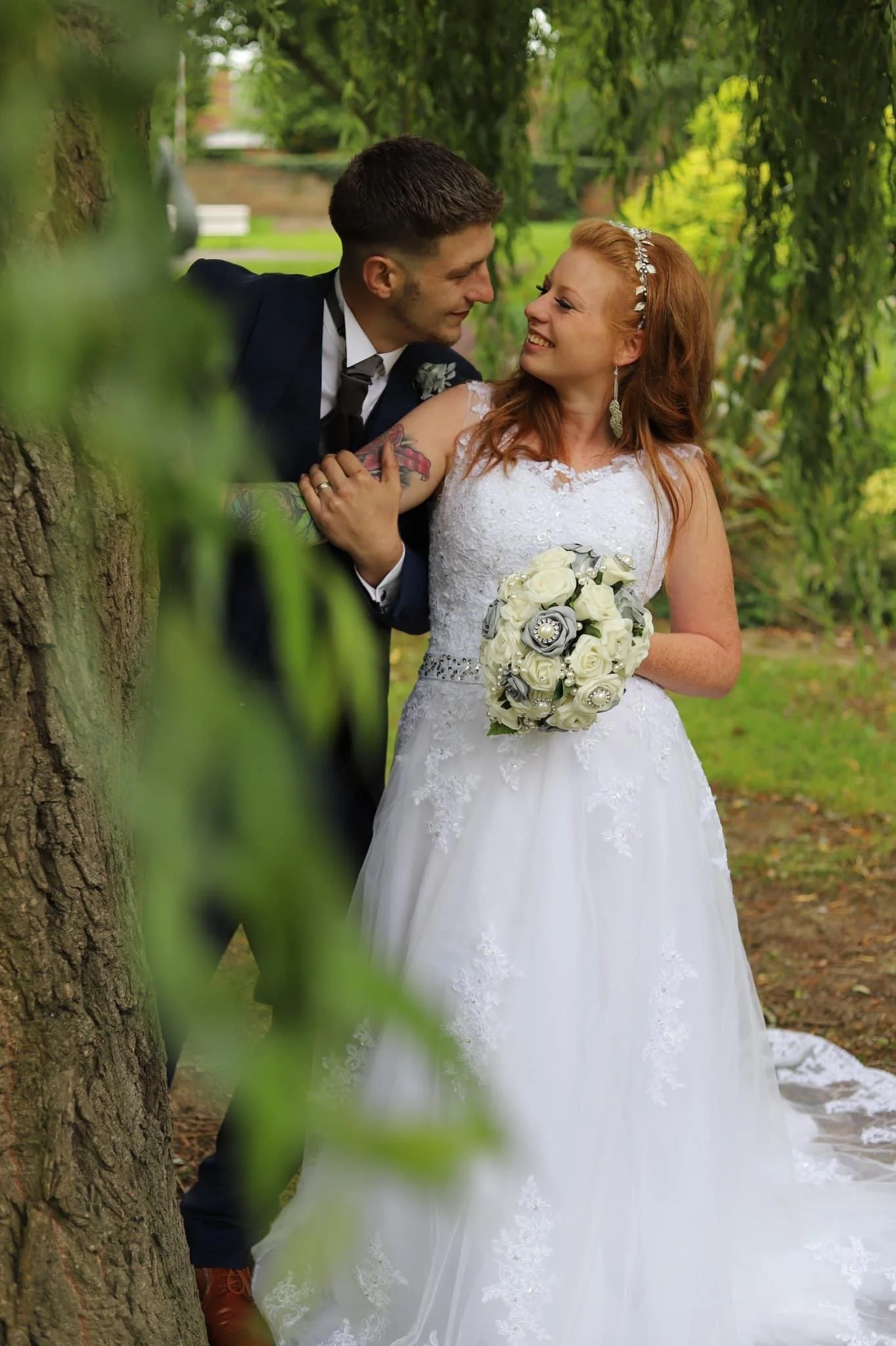A bride and groom in wedding attire stand outdoors in a lush green park, sharing a joyful moment with the bride holding a bouquet of white roses and silver decorations.