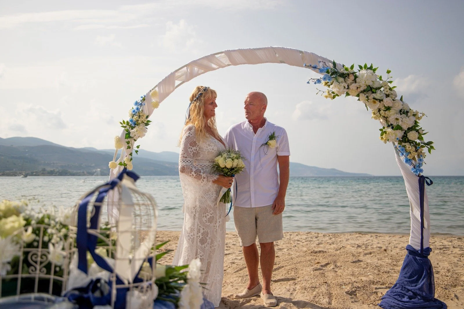 A bride and groom stand under a floral arch on a sandy beach, facing each other during their wedding ceremony, with the ocean and mountains in the background.