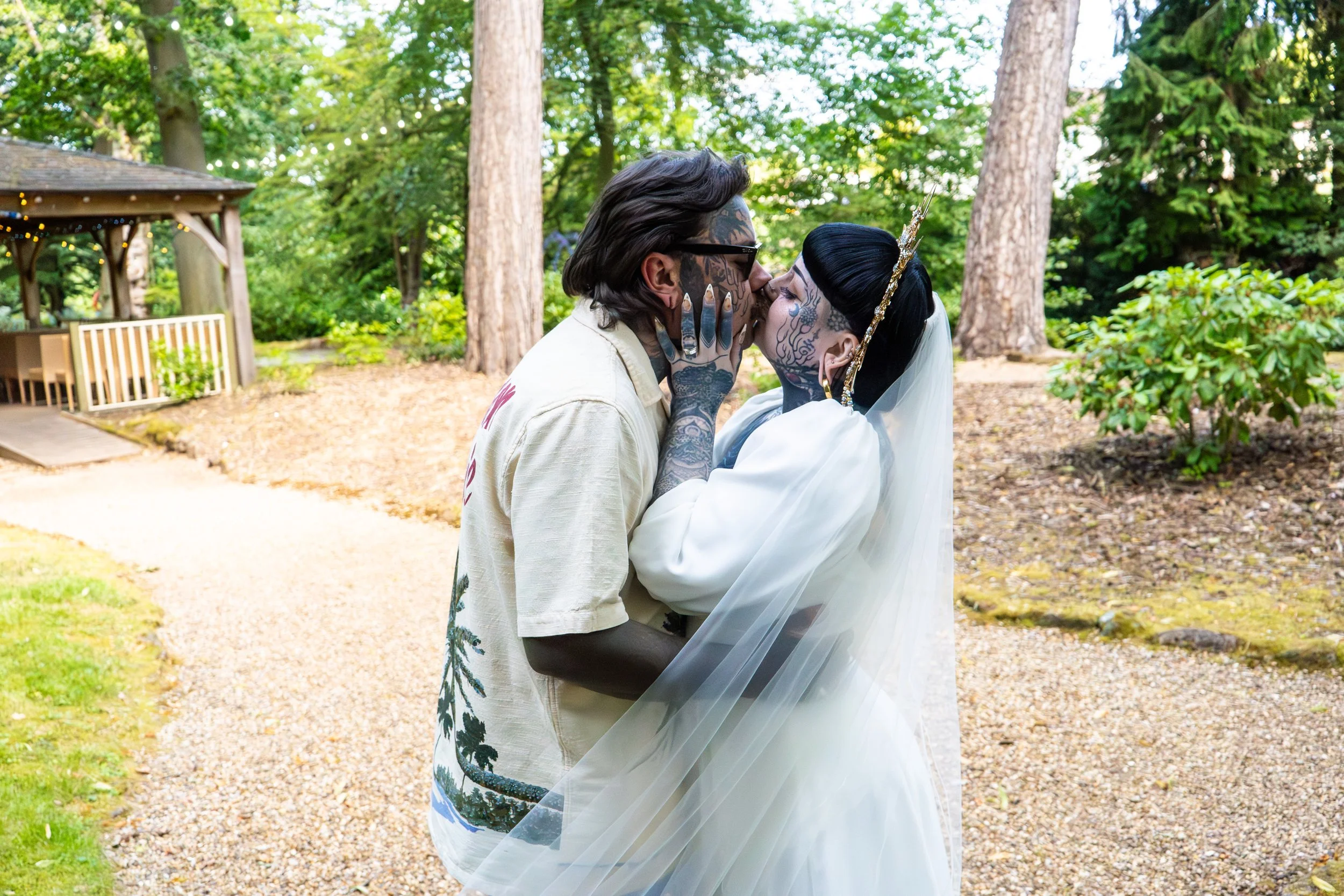 A couple dressed in wedding attire kissing outdoors in a wooded area with trees and greenery.