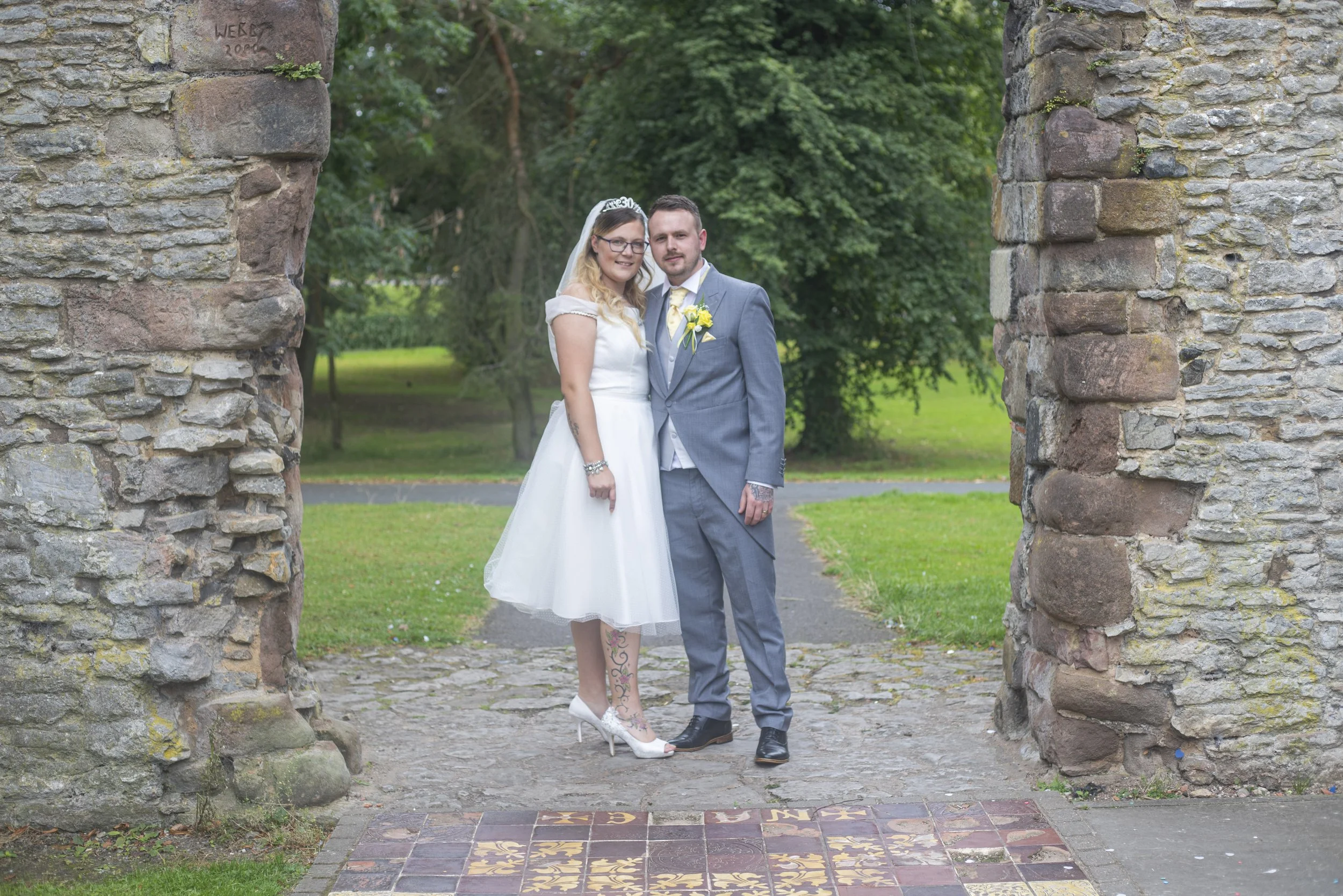 A bride and groom standing together outdoors on a wedding day, framed by stone archways with green trees and grass in the background.