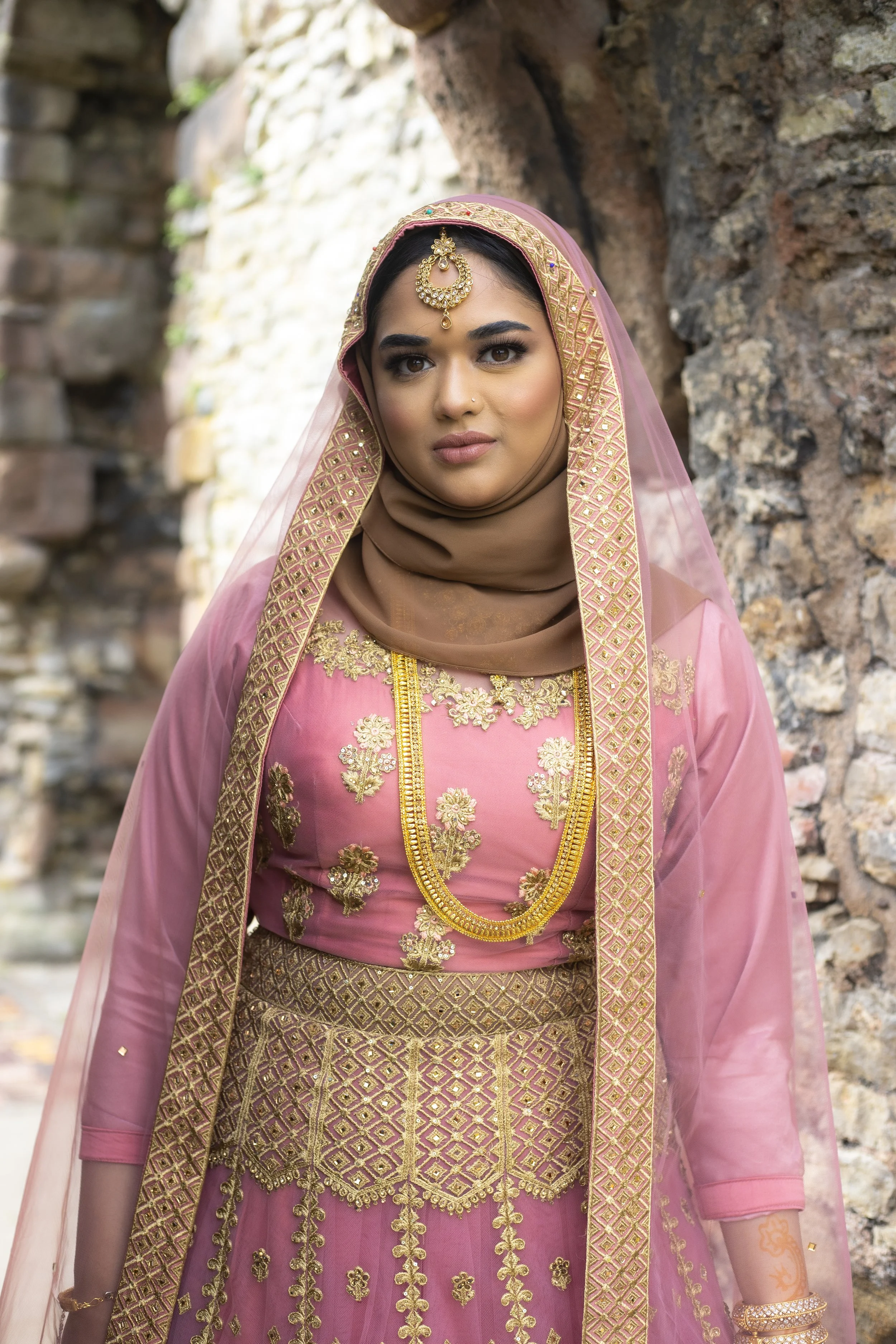 A woman dressed in traditional Indian pink and gold attire standing outdoors near a stone wall.