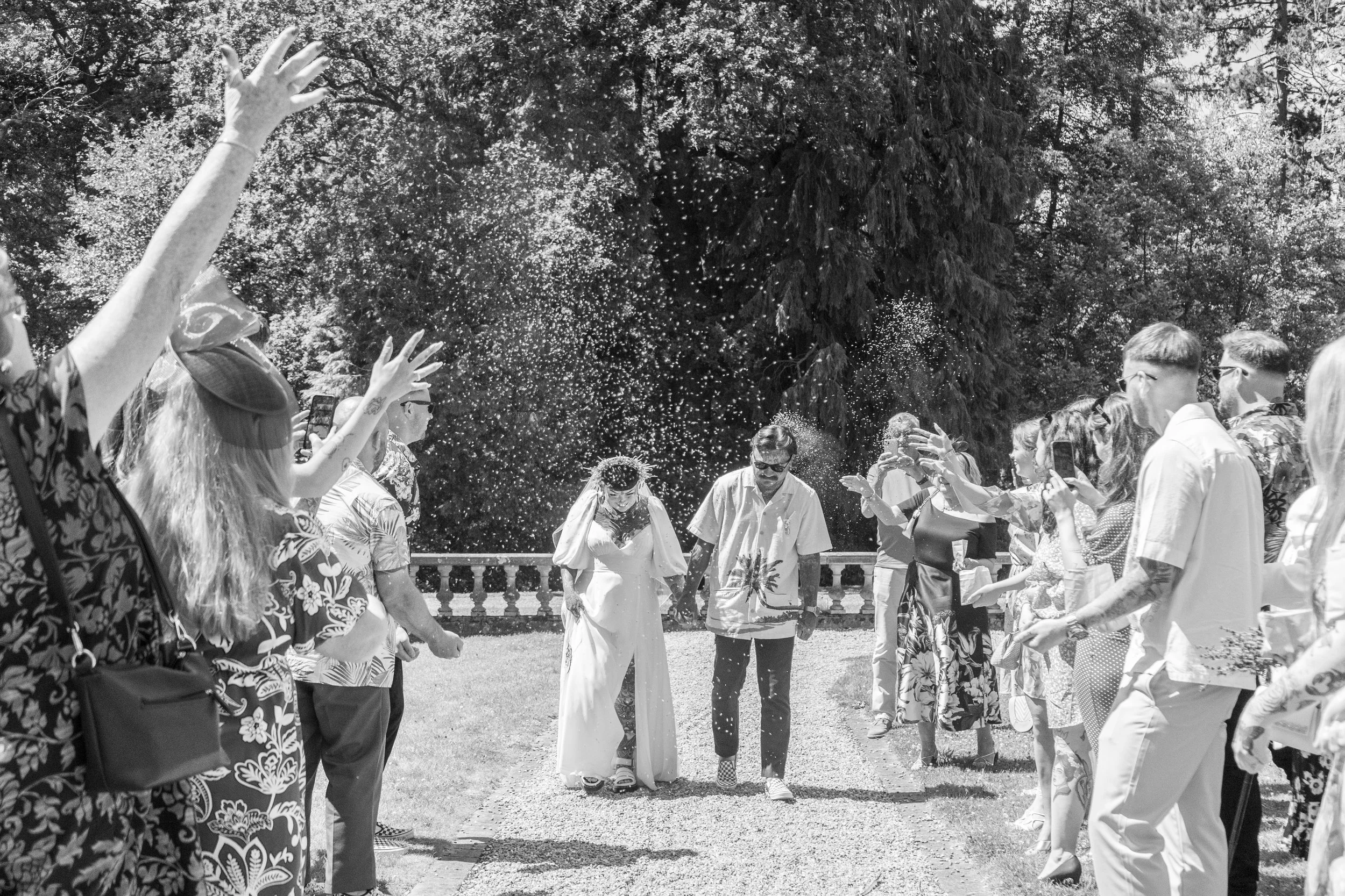 Black and white photo captures a wedding send-off with drops of water or confetti in the air. The bride and groom are walking arm-in-arm, surrounded by guests on either side who are throwing celebratory items and taking photos outdoors.