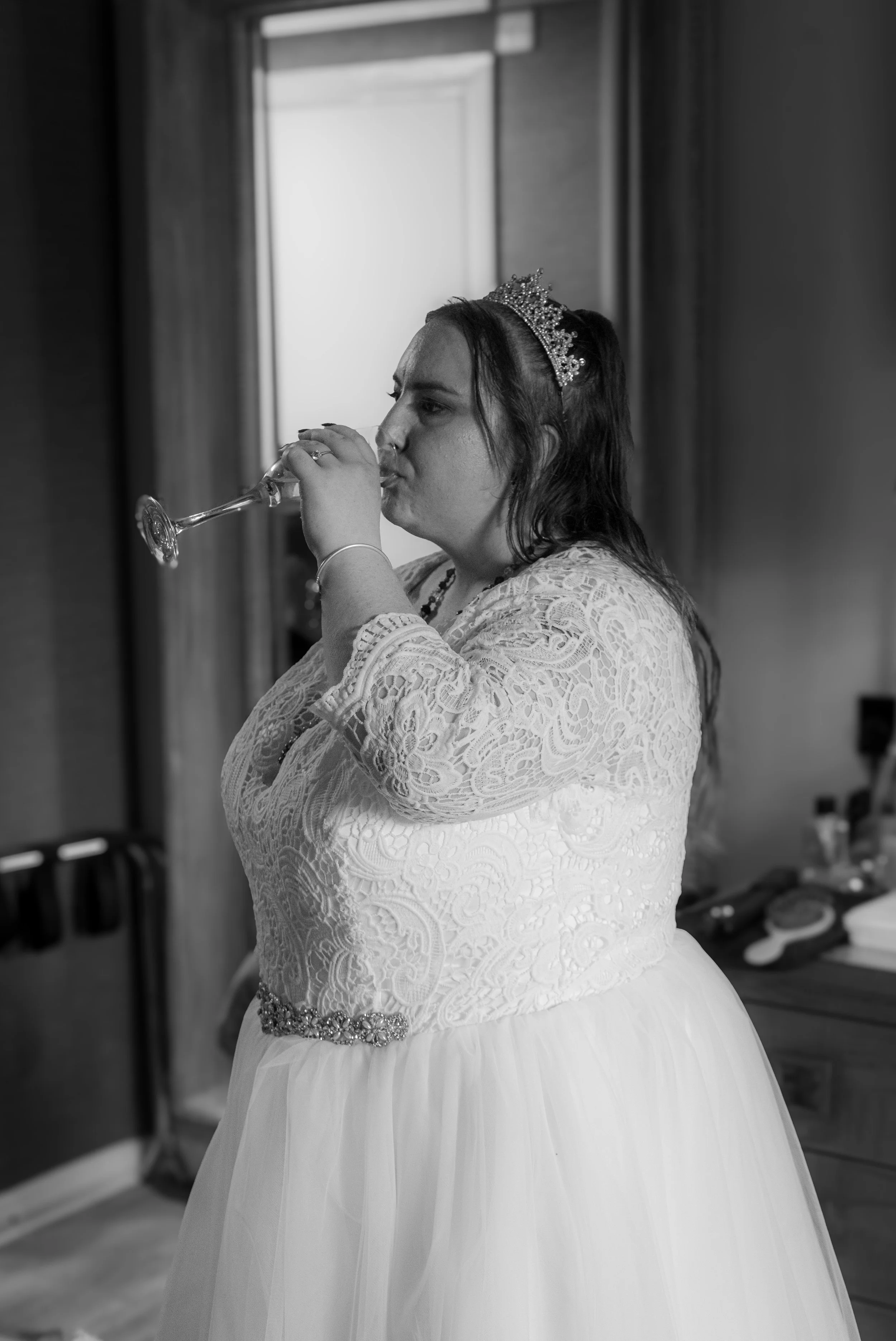 A woman in a wedding dress and tiara drinks champagne in a room with curtains and a dresser.