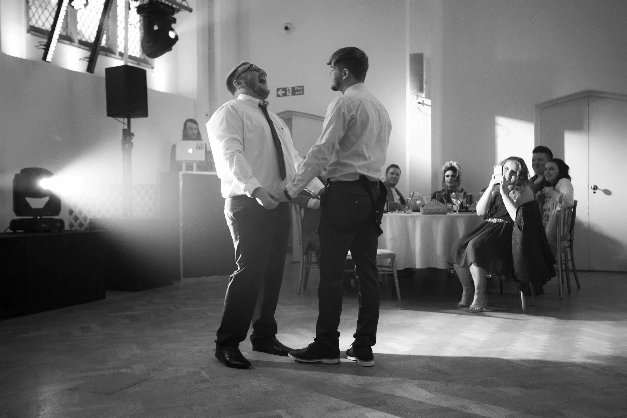Two men holding hands and smiling while dancing at a wedding reception, with guests watching and taking photos in the background.