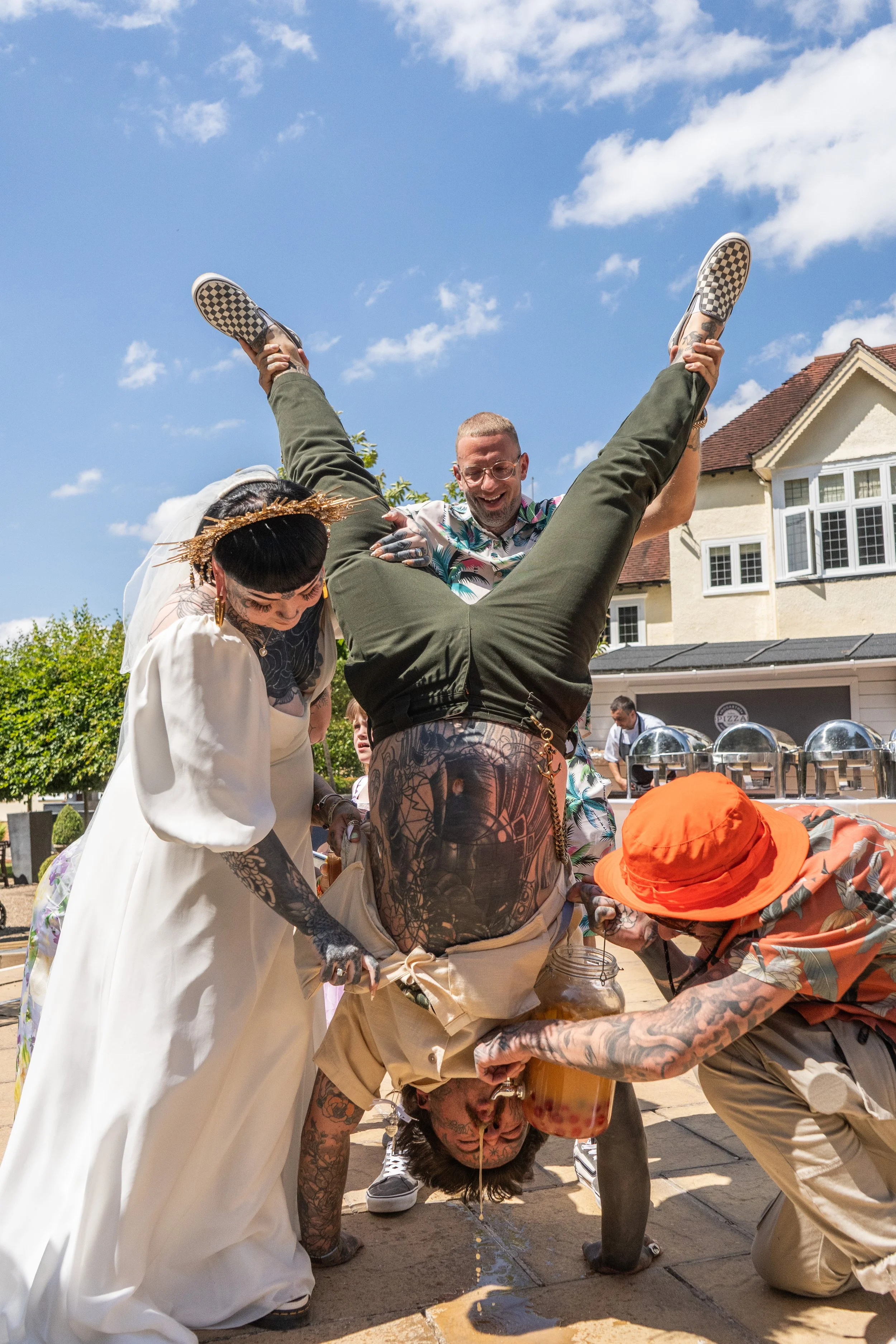 Group of people participating in a playful outdoor event, with one person upside down, others assisting, under a partly cloudy sky.