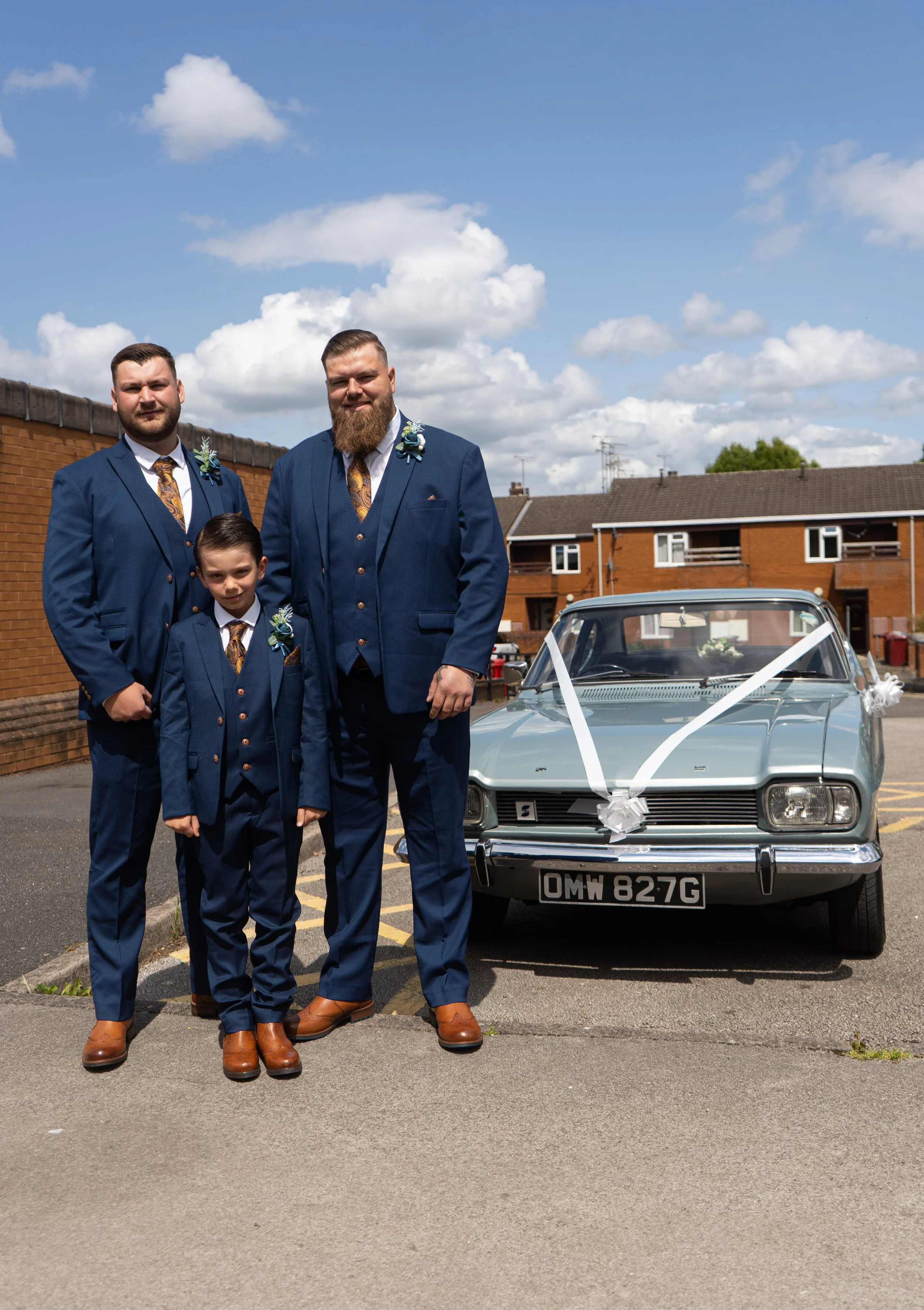 Three men and one boy dressed in blue suits with brown shoes standing next to a vintage gray car decorated with white ribbons, set outdoors on a sunny day with clouds and a residential area in the background.