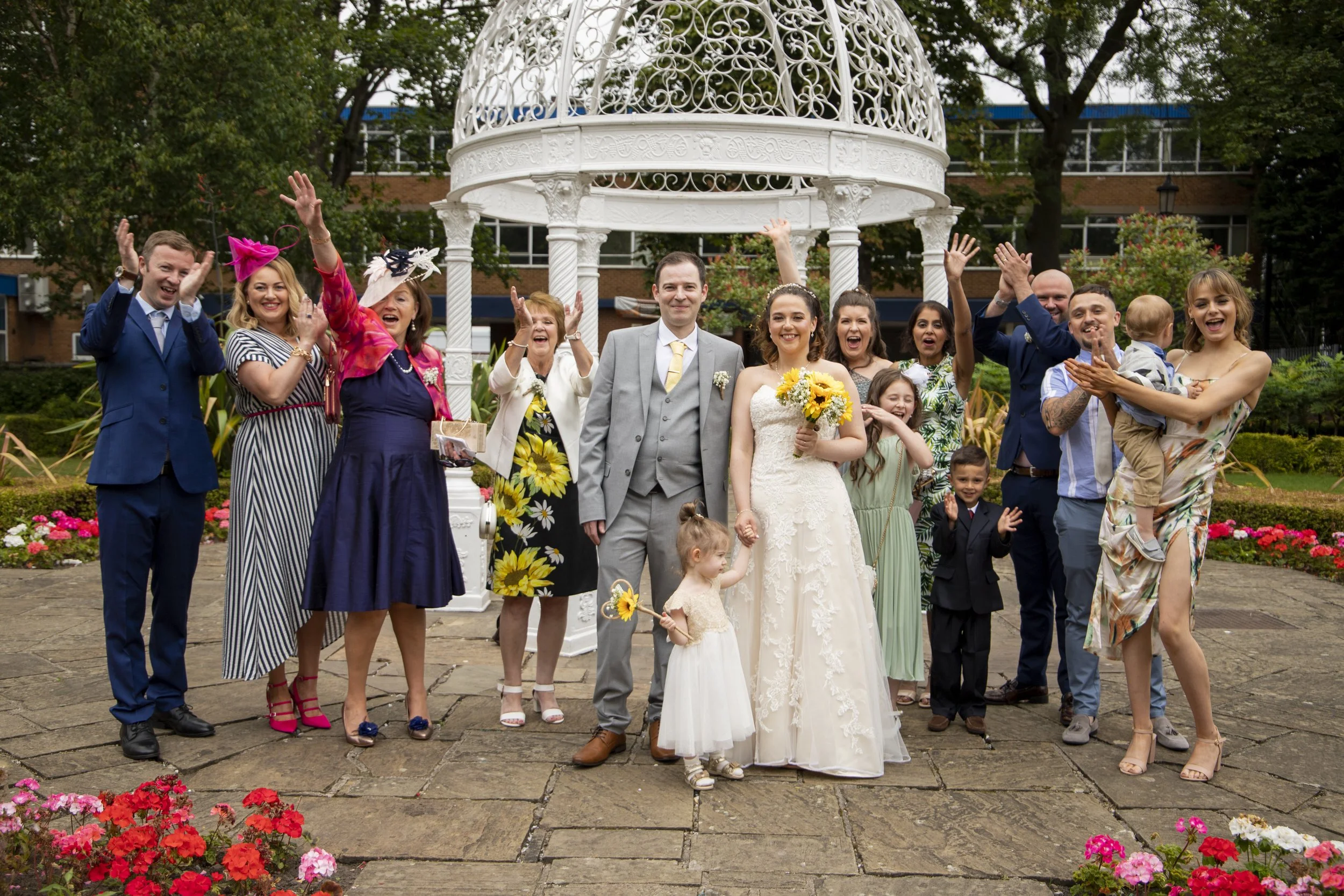 A group of people, including a bride and groom, celebrating outdoors on a brick patio with flowers and trees in the background, under a decorative white gazebo.