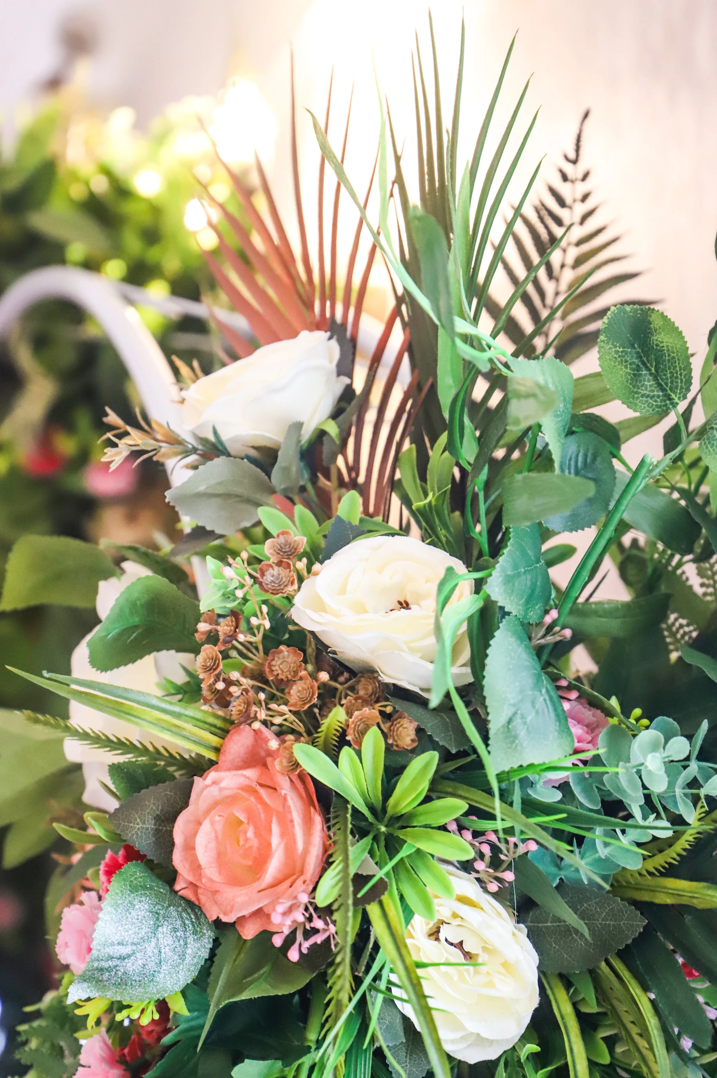 Close-up of a colorful artificial flower bouquet with white, pink, and peach roses surrounded by various green leaves and ferns, with sunlight in the background.