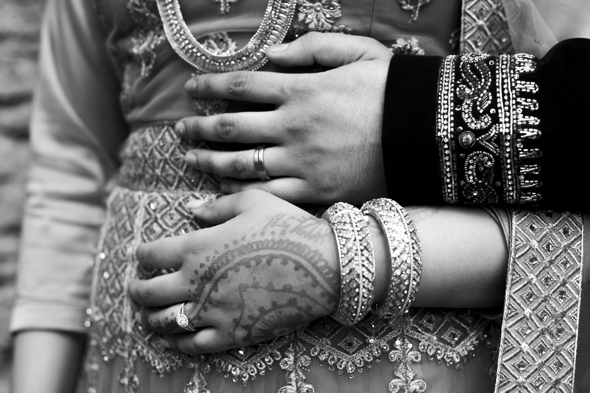 Close-up of a woman's hand resting on her chest, wearing ornate jewelry including bangles, rings, and a necklace, with intricate embroidery on her traditional dress. The image is in black and white.