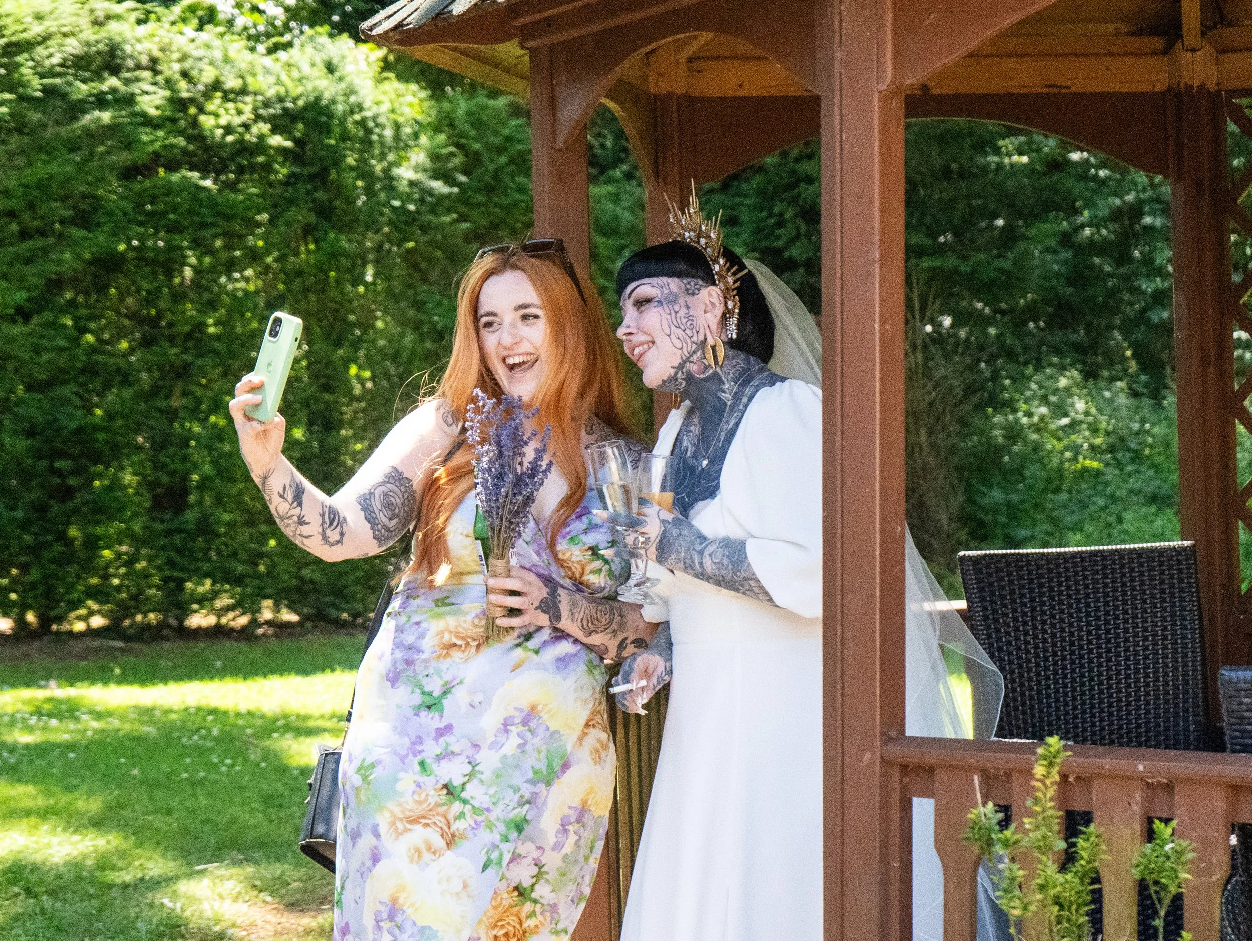 Two women, one with red hair and tattoos wearing a floral dress, taking a selfie with a woman wearing a wedding dress and tattoos, holding glasses of champagne, outdoors on a sunny day near a wooden gazebo.