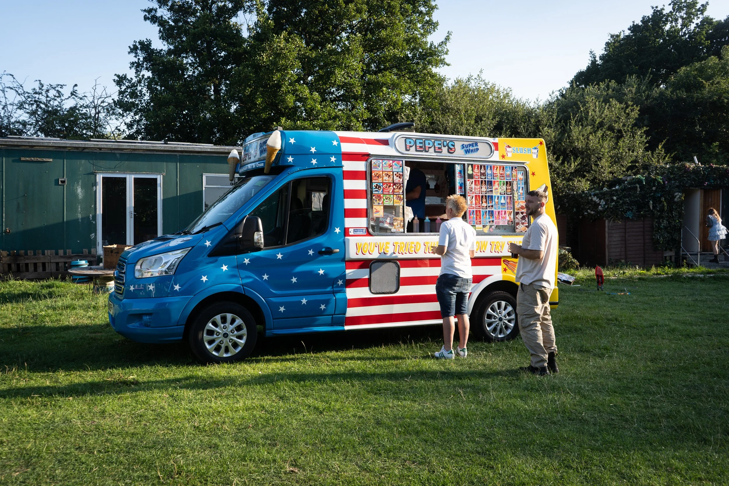 A colorful ice cream truck decorated with an American flag motif on a grassy area, with two people standing in front of it and a woman walking toward a small shed in the background.