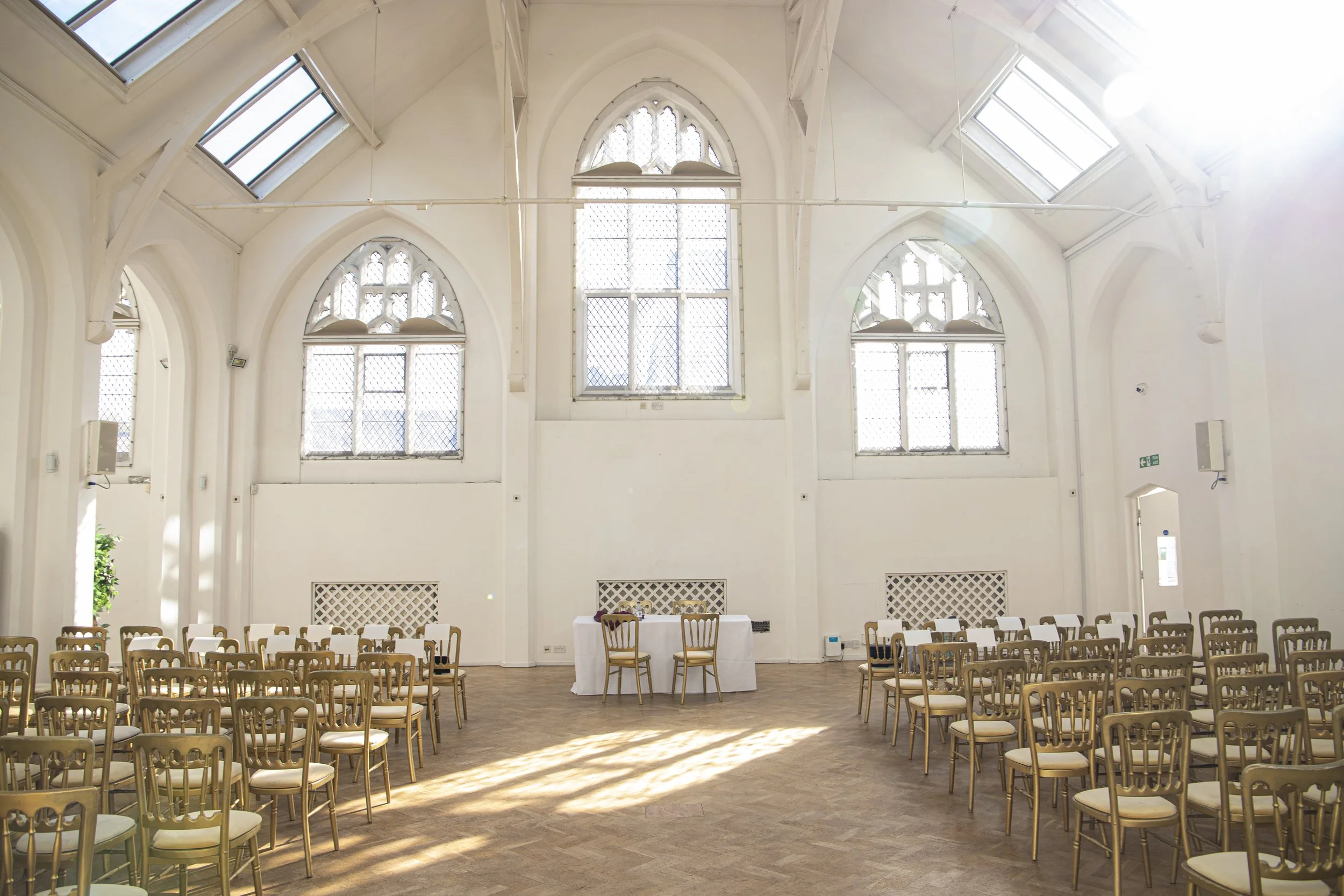 An interior of a bright, spacious room with high arched ceilings, large windows, and multiple rows of chairs arranged for an event. The room has wooden flooring and is illuminated by natural sunlight.