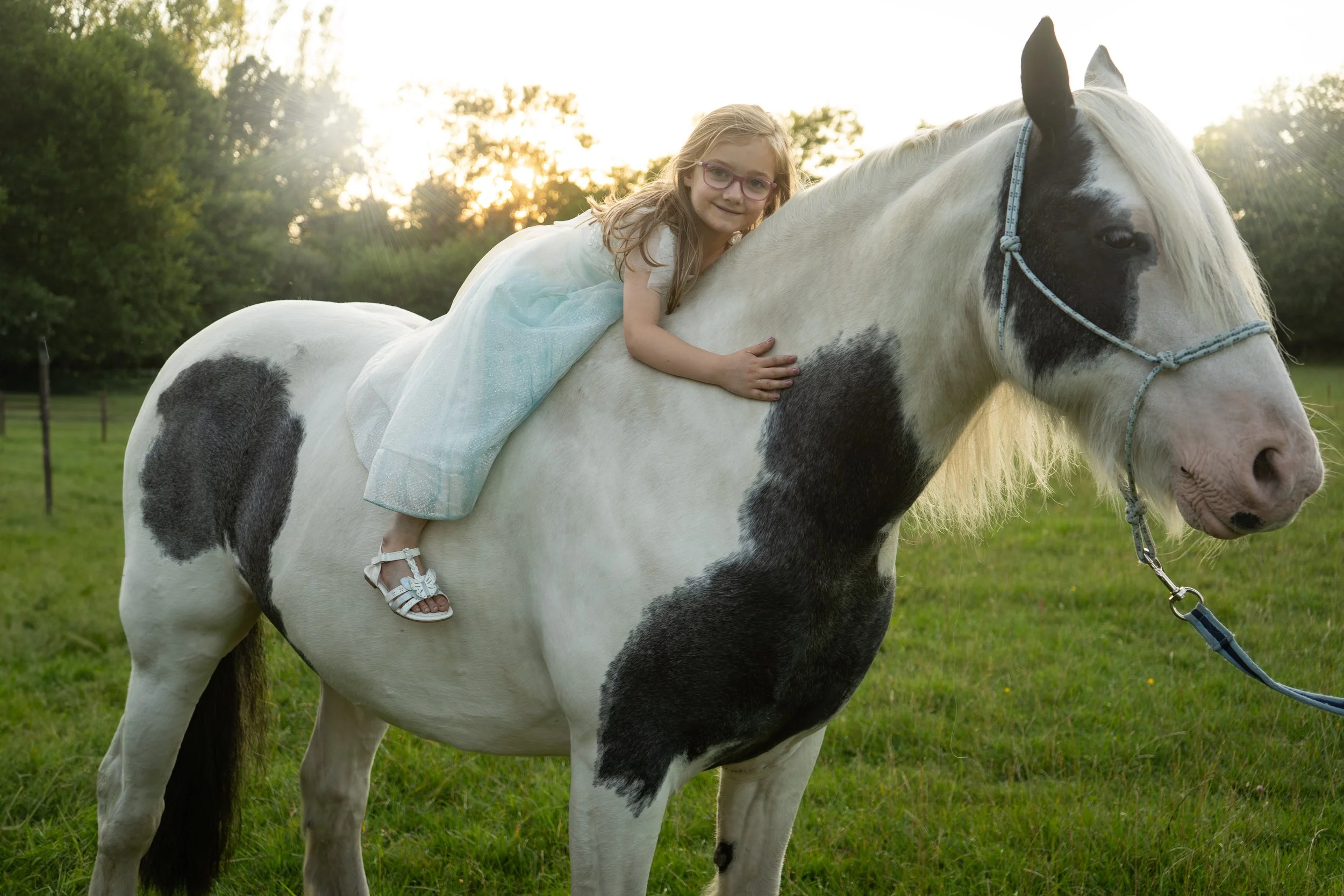A young girl with glasses and a light dress riding a white and black spotted horse in a grassy field with trees and sunset in the background.