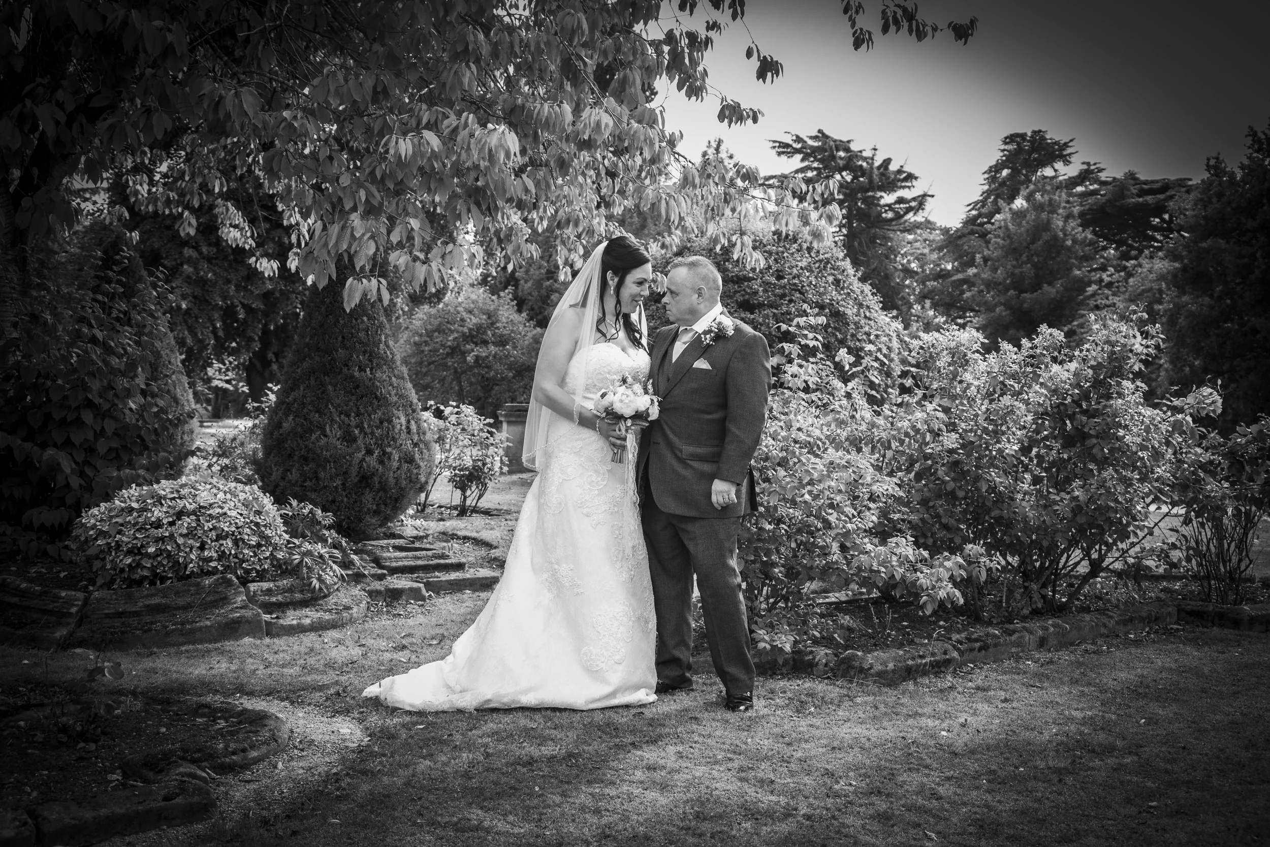 A black and white photo of a bride and groom standing close in a garden, gazing into each other's eyes. The bride is holding a bouquet and wearing a lace wedding dress and veil. The groom is in a suit with a boutonniere. There are trees and bushes in the background.
