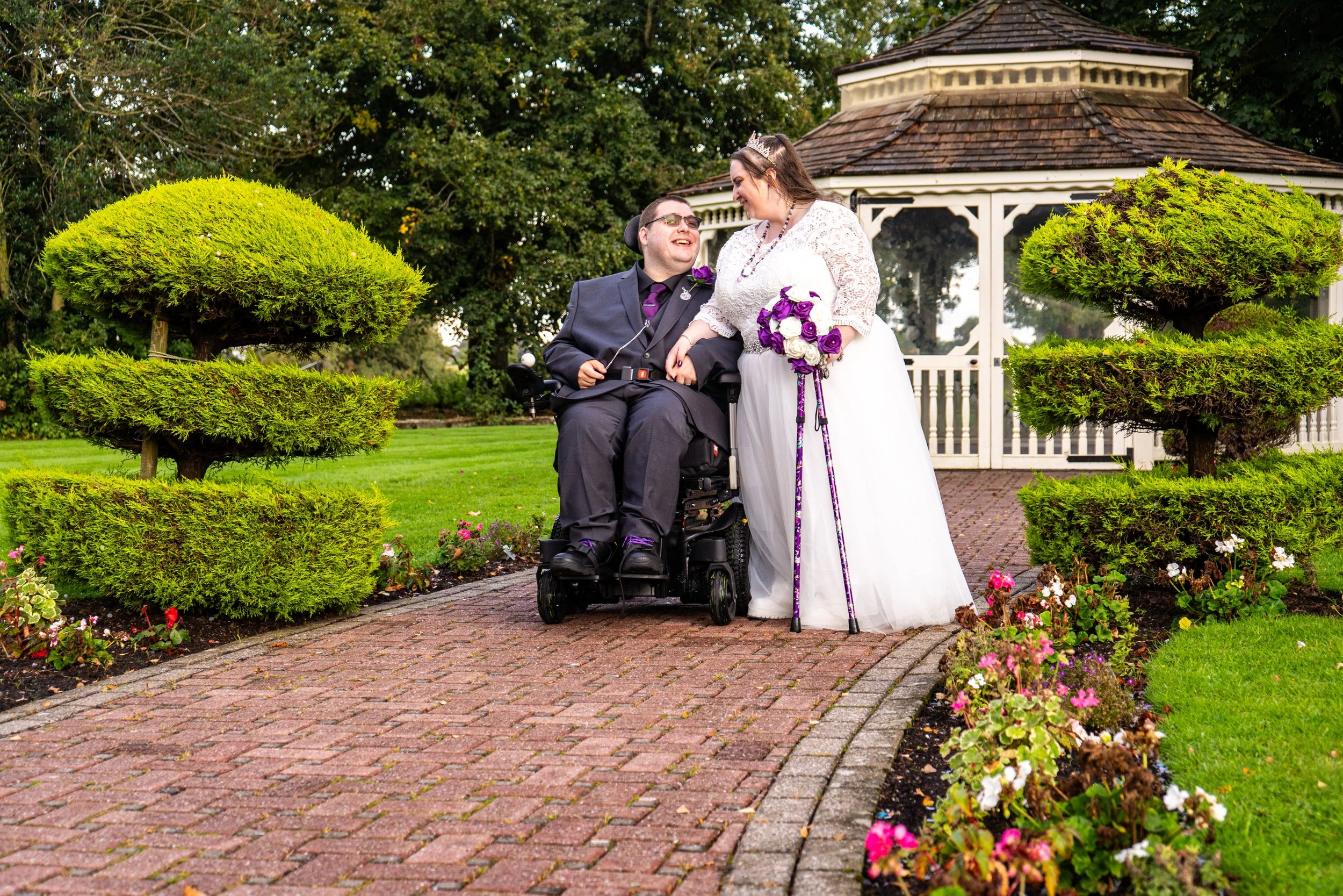 Couple on wedding day, one in wheelchair, sharing a moment outdoors near a gazebo, surrounded by well-manicured garden with green shrubs and flowers.