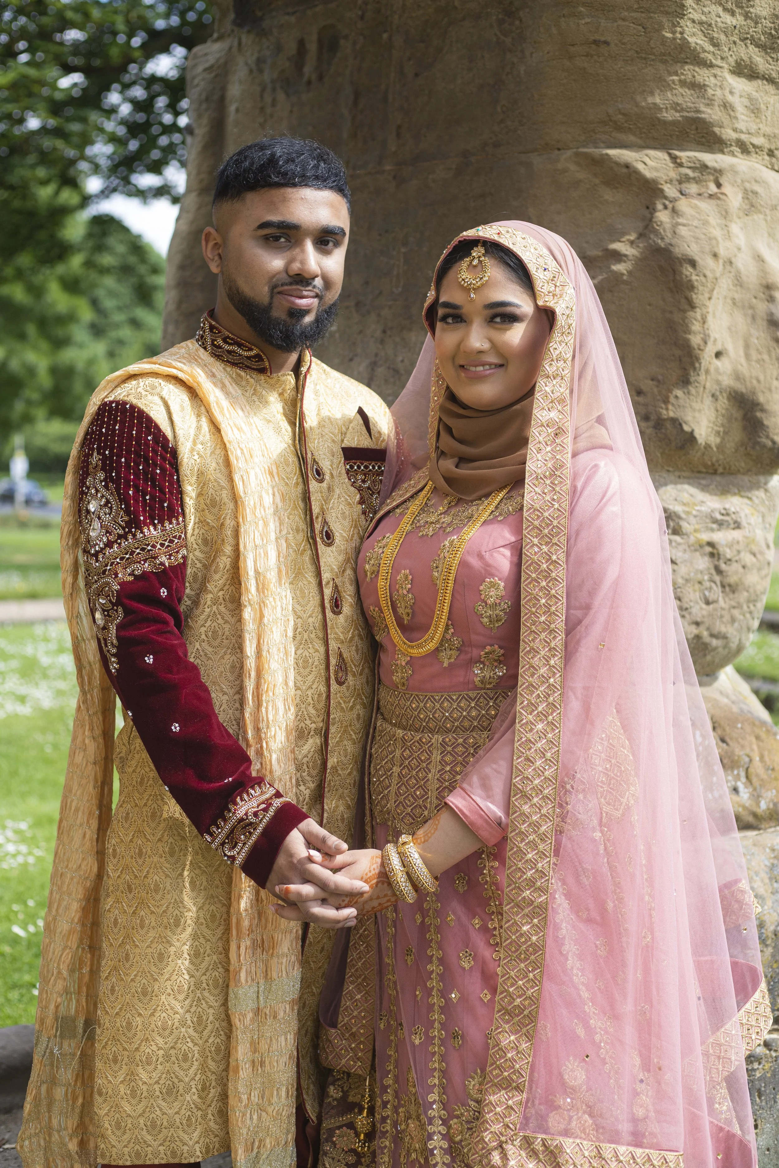 A couple dressed in traditional South Asian wedding attire holding hands outdoors, standing in front of a large stone structure with greenery in the background.