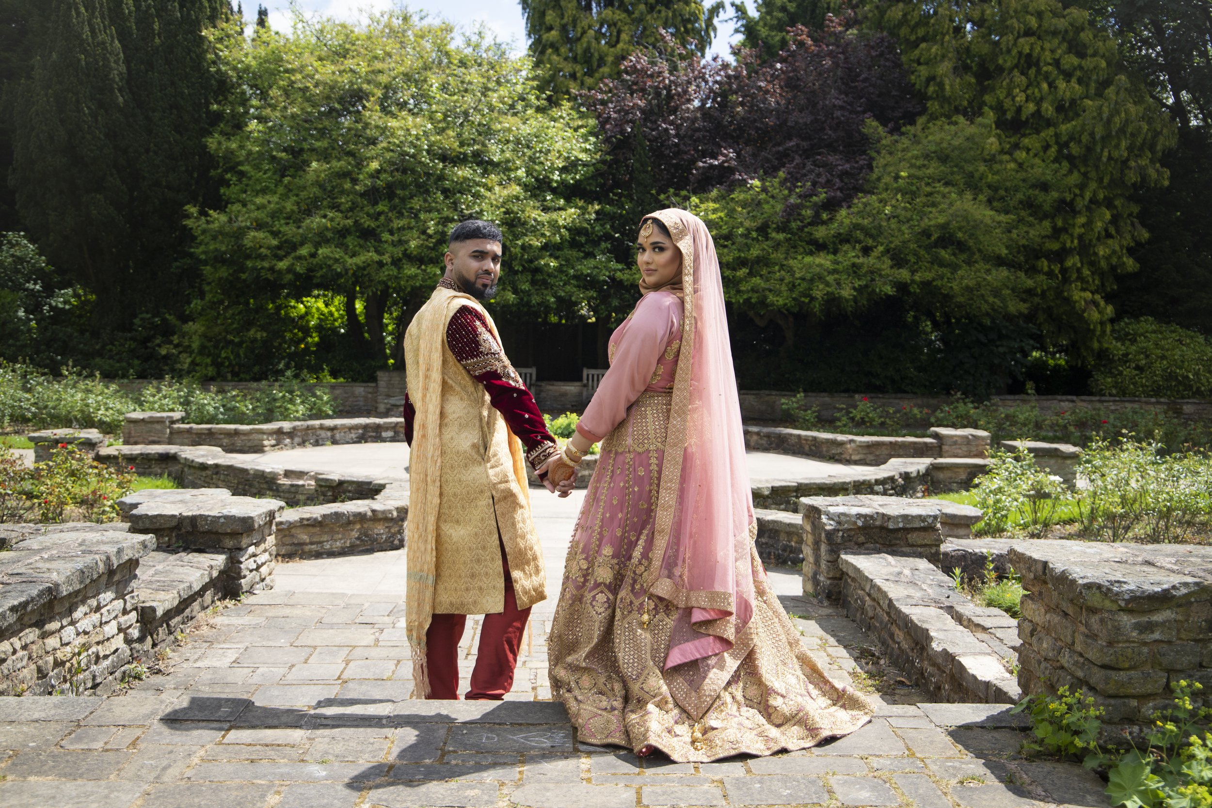 A couple dressed in traditional South Asian wedding attire standing hand in hand in a garden with lush green trees and stone pathways.