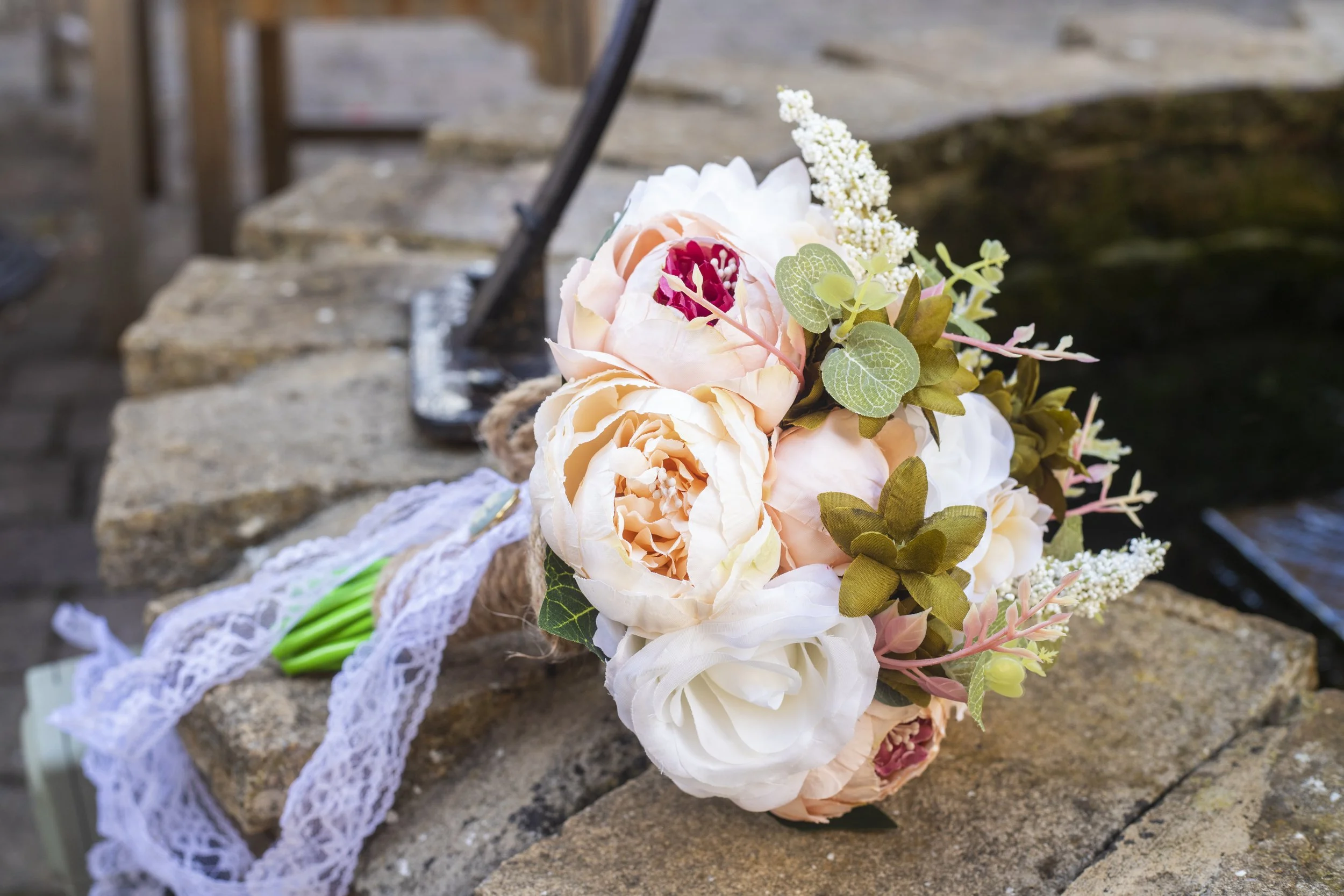 A bouquet of peach and white flowers with green leaves and white lace ribbon, resting on a stone surface outdoors.
