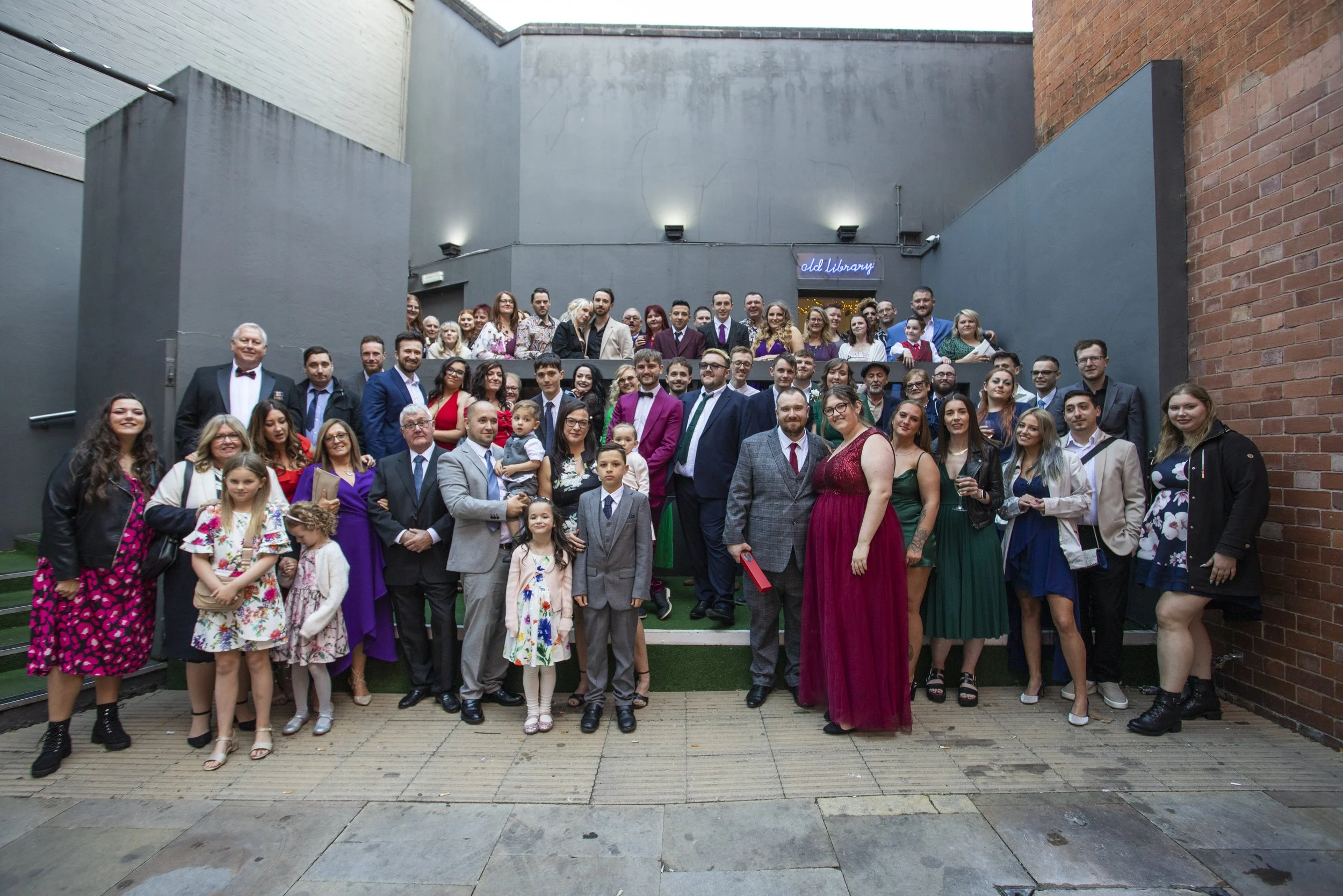 A large group of people dressed in formal and semi-formal attire, gathered outdoors on a small set of stairs and the surrounding pavement, for a group photo at a celebration or event, with a sign that reads 'old library' in the background.