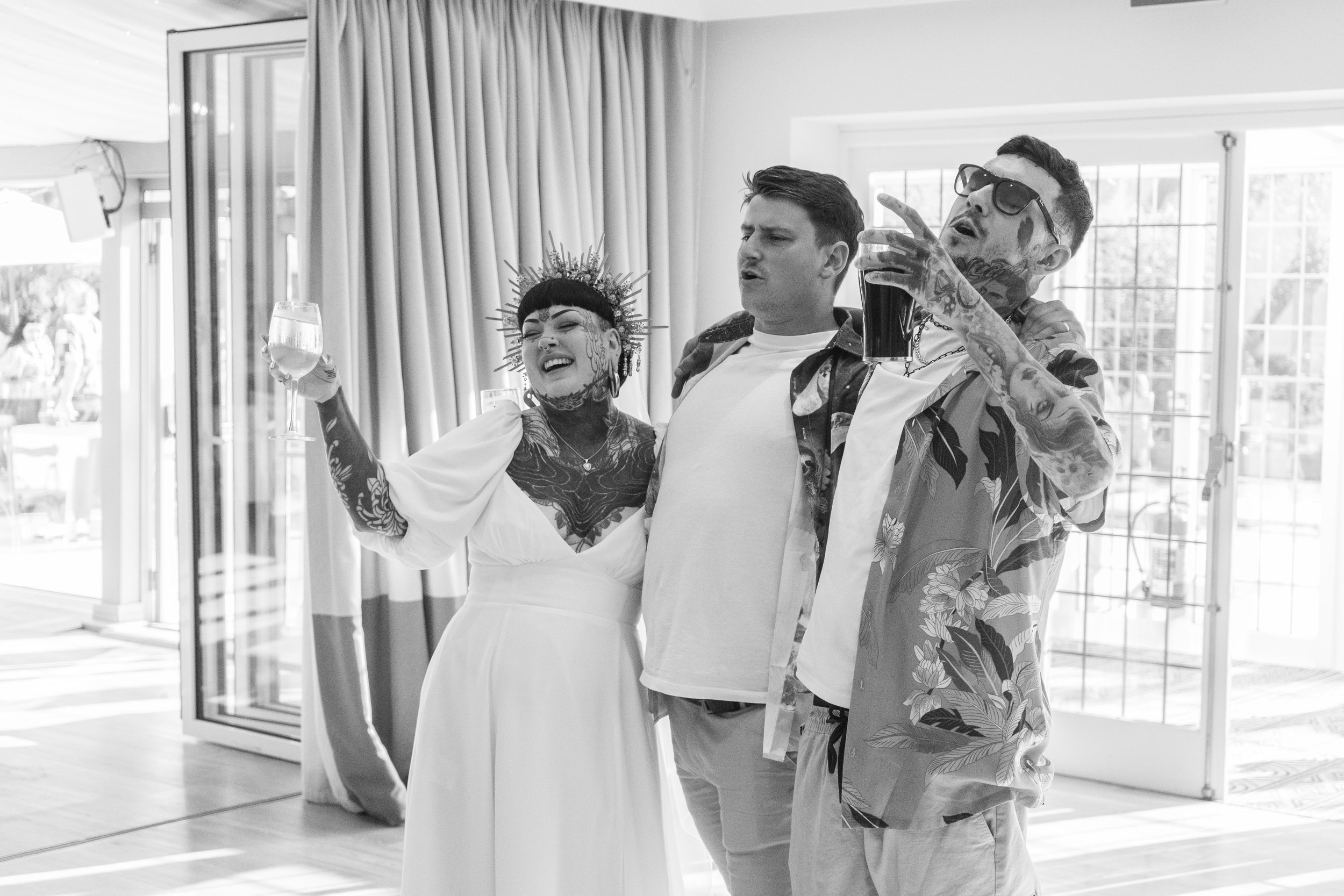 Three people with tattoos and tattoos, one woman wearing a crown and a white dress, two men holding drinks, celebrating indoors with open sliding doors and curtains.