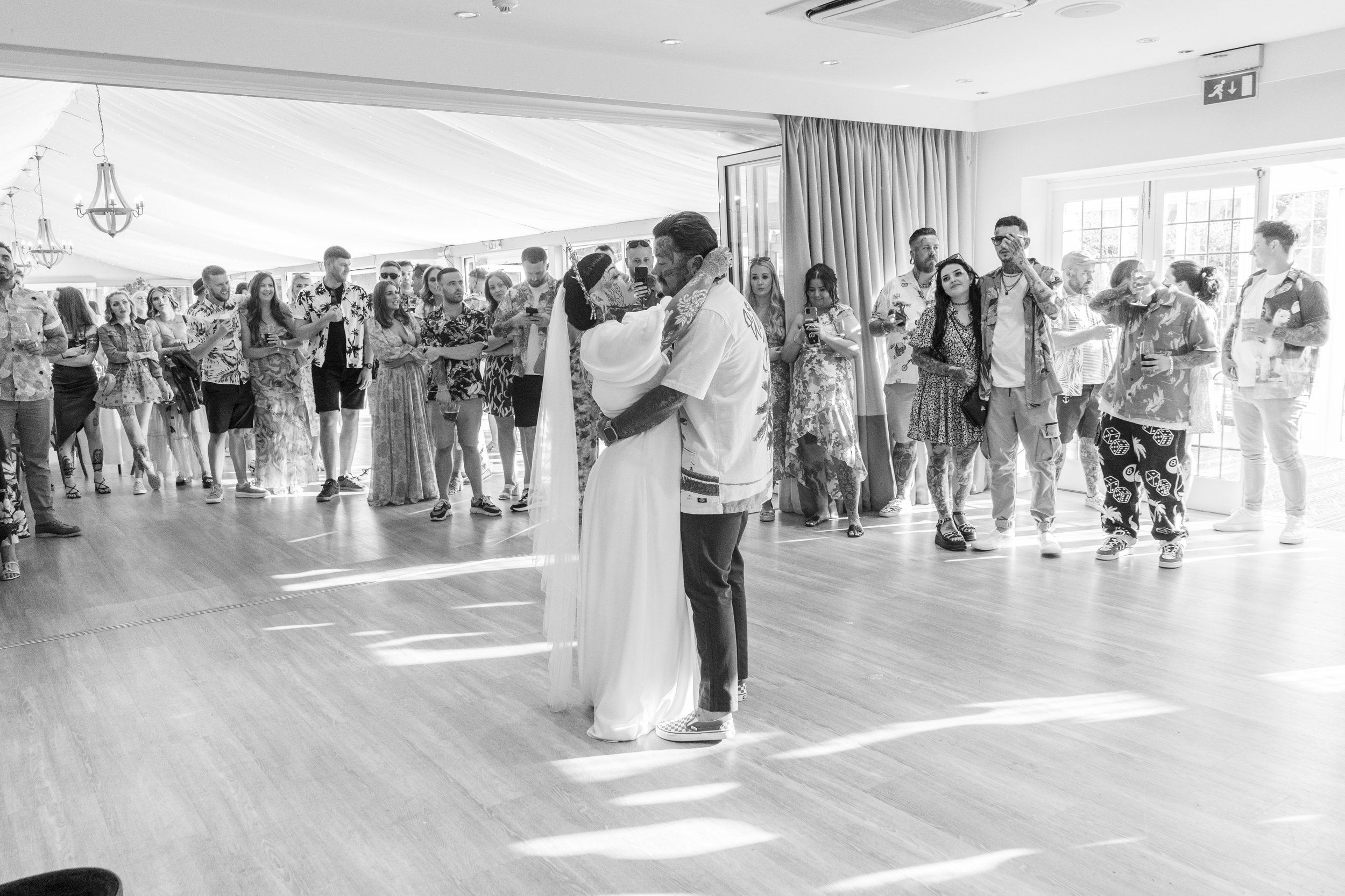 A bride and groom sharing a dance at their wedding reception, surrounded by guests in tropical-themed clothing, in a decorated indoor venue with sunlight coming through windows.