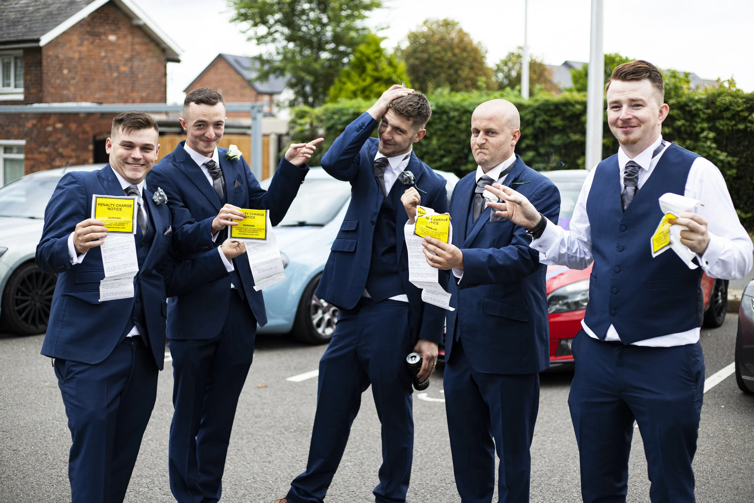 Five men in suits stand outside, holding parking violation notices, with playful expressions. One is touching his head, and others are smiling or making gestures, standing in a parking lot with cars and houses in the background.