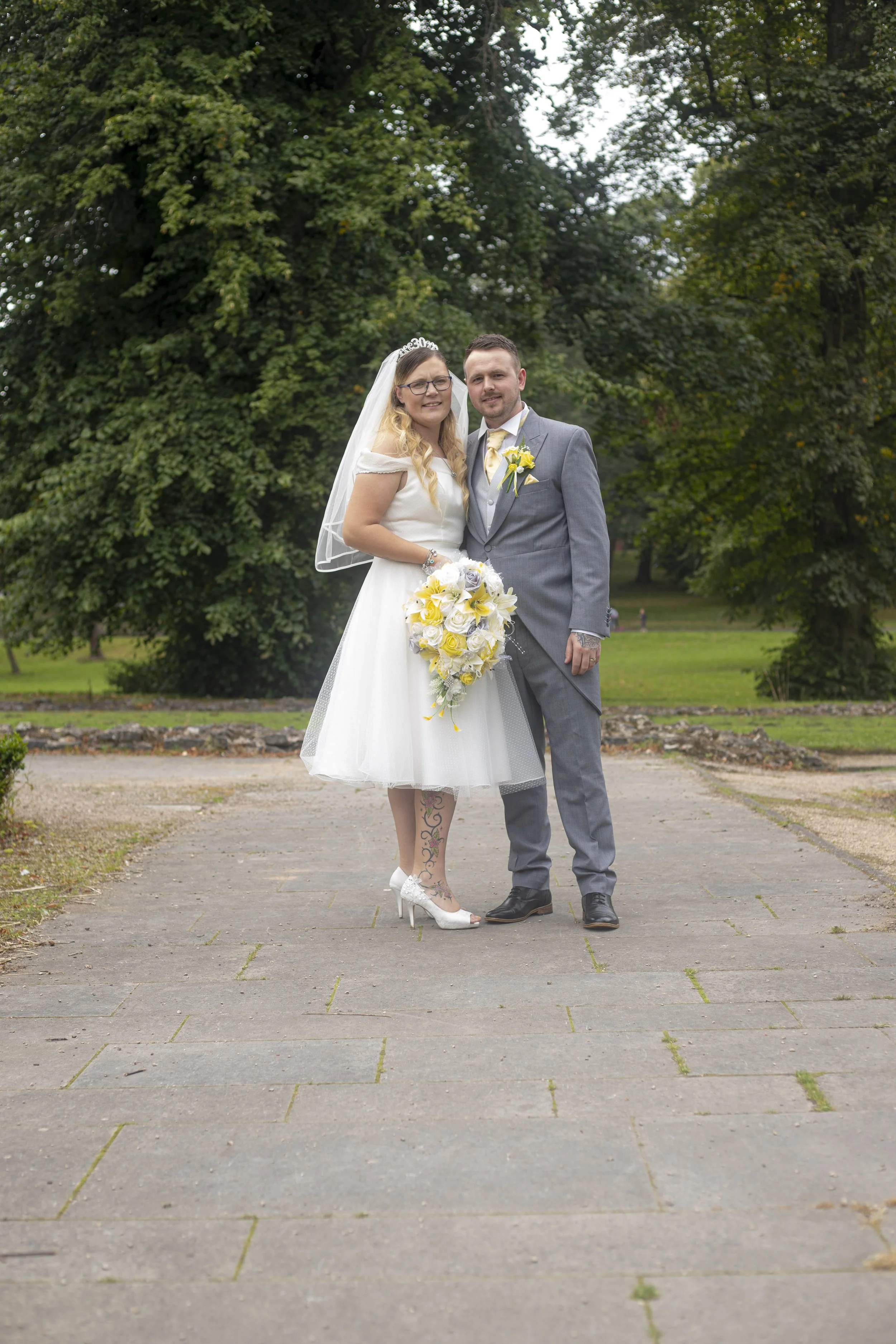 A wedding couple standing outdoors on a paved pathway, surrounded by green trees, with the bride holding a large bouquet of yellow and white flowers.