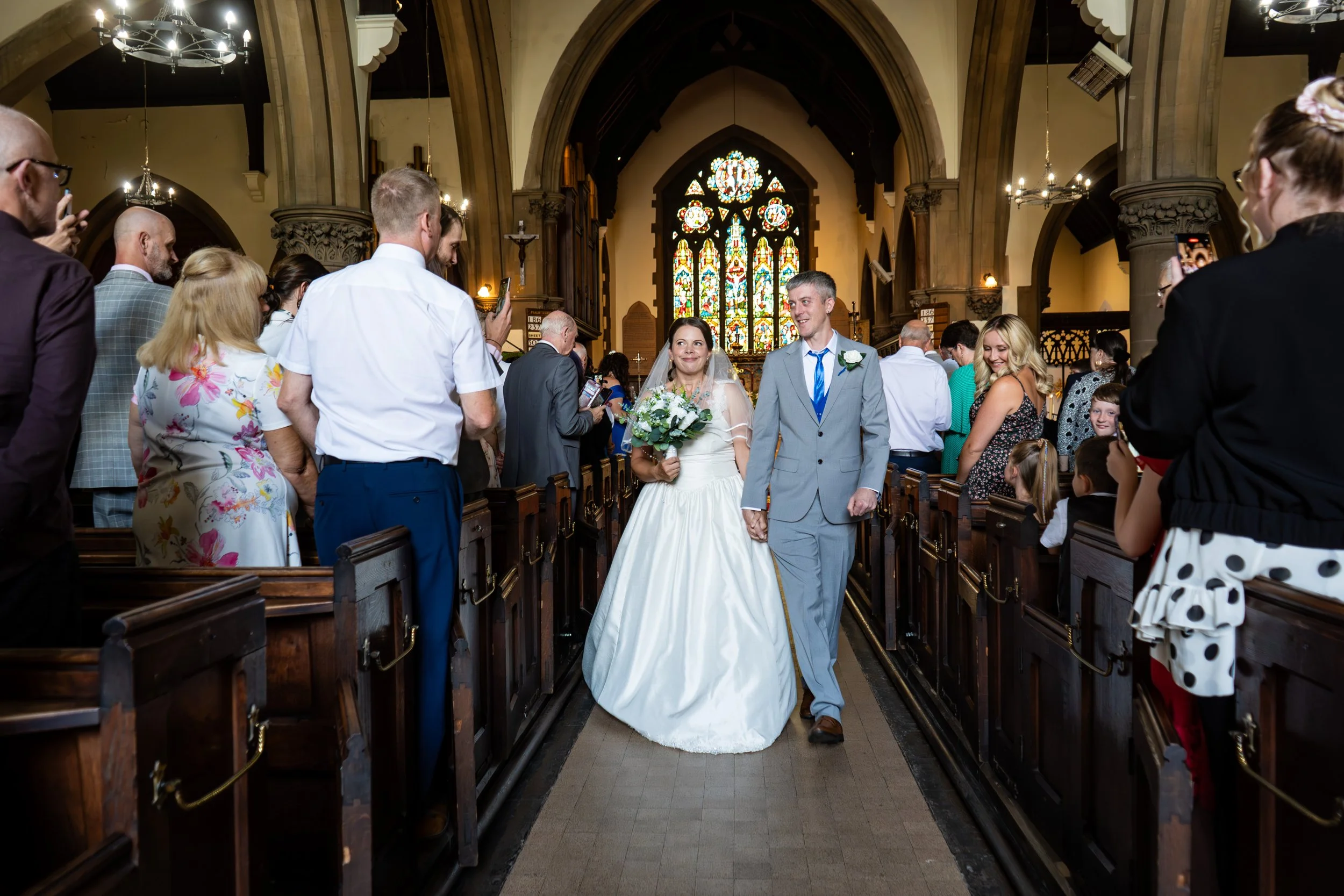 A bride and groom walking down the aisle of a church after their wedding ceremony, surrounded by family and friends, with stained glass windows in the background.