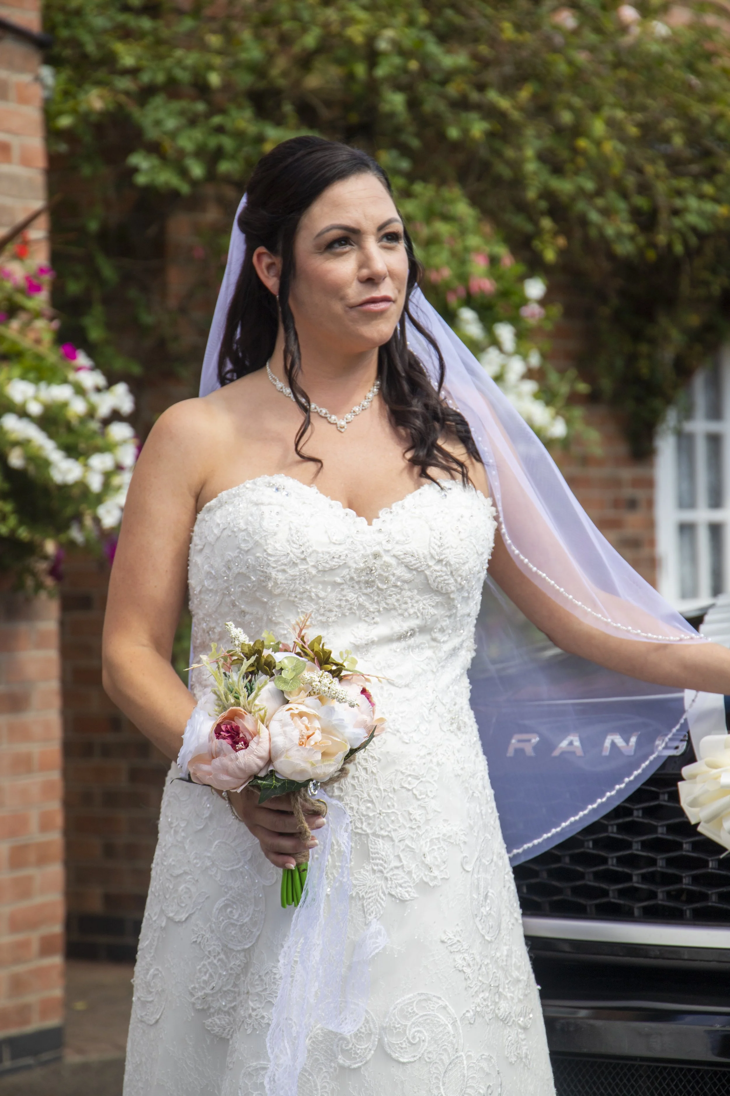 A bride in a white lace wedding dress holding a bouquet of pastel flowers, standing outdoors with a brick wall and green foliage in the background.