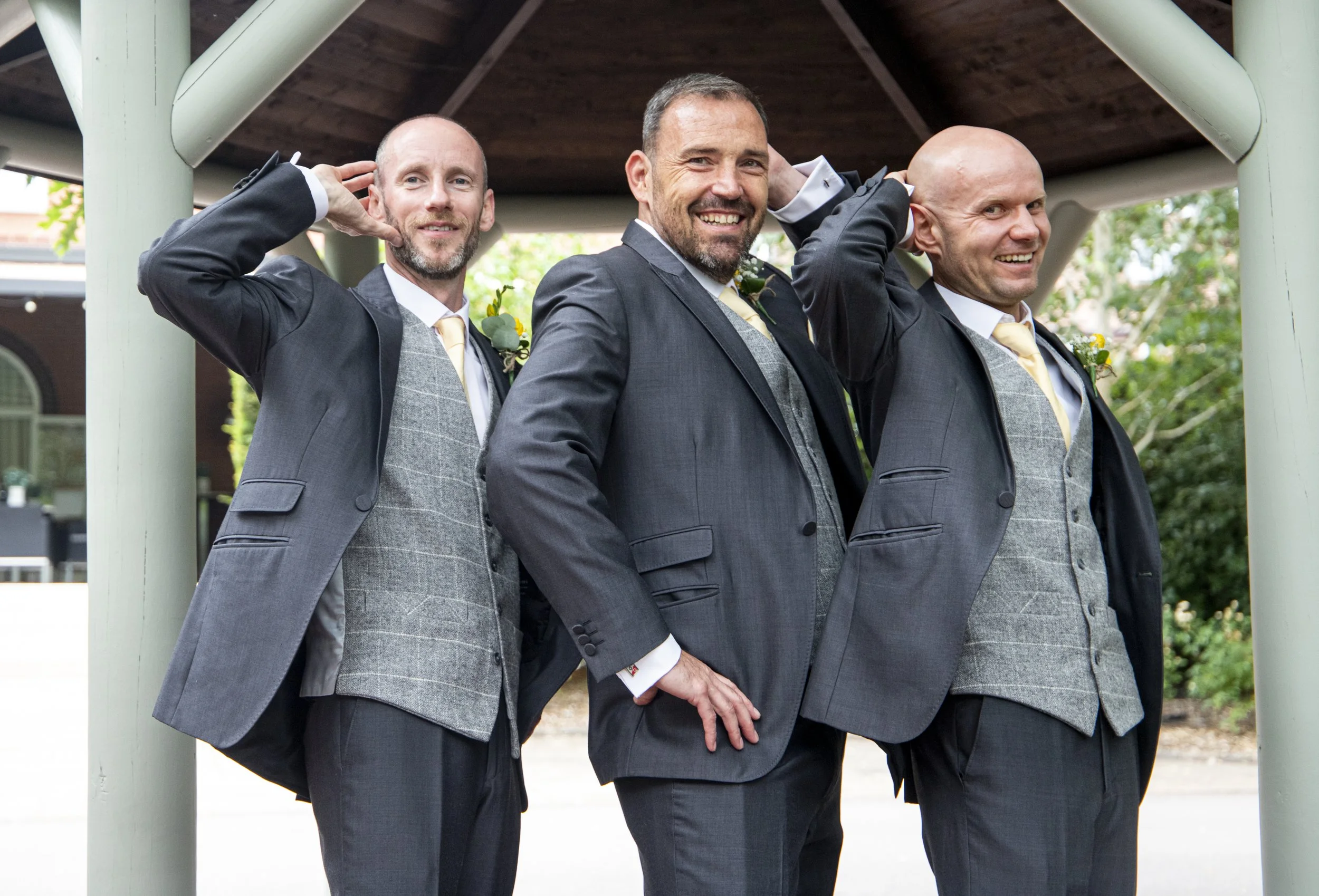 Three men in suits standing under a gazebo, smiling and adjusting their ears, with one man in the middle and the other two on either side.