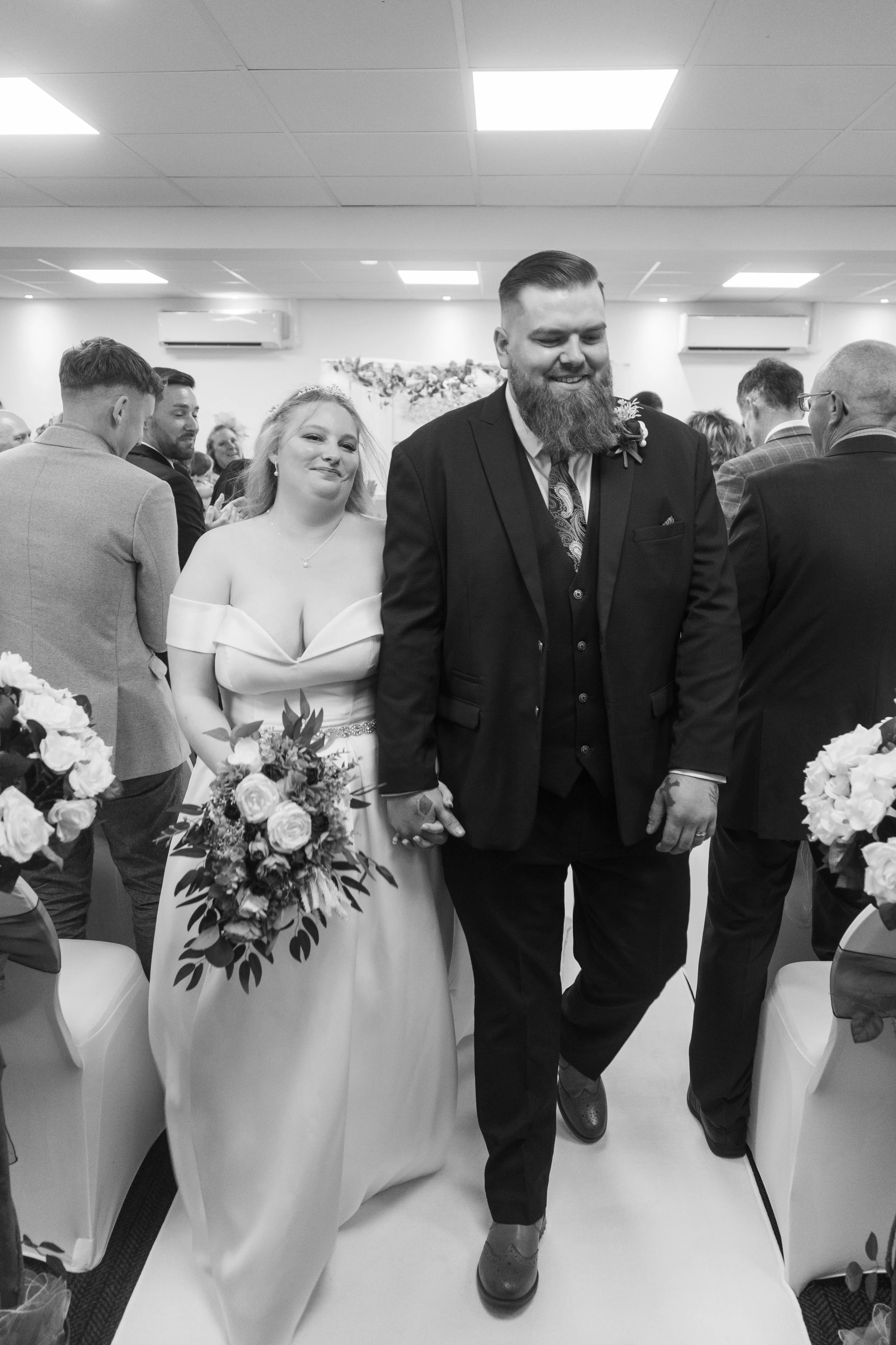 A black and white photo of a bride and groom holding hands and walking down the aisle, surrounded by wedding guests in an indoor venue.