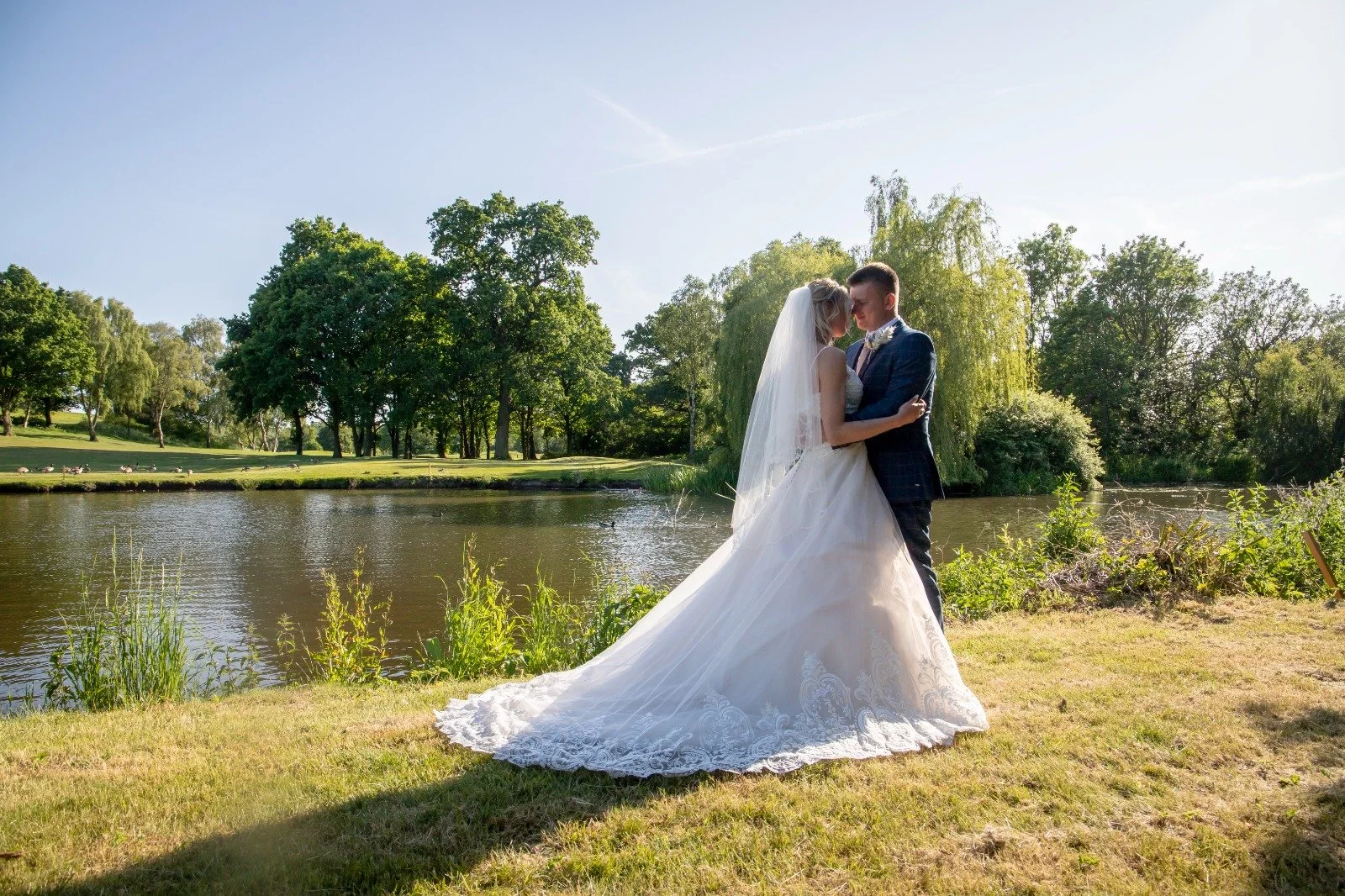 A bride and groom embrace near a lake in a park with trees, under a clear sky.