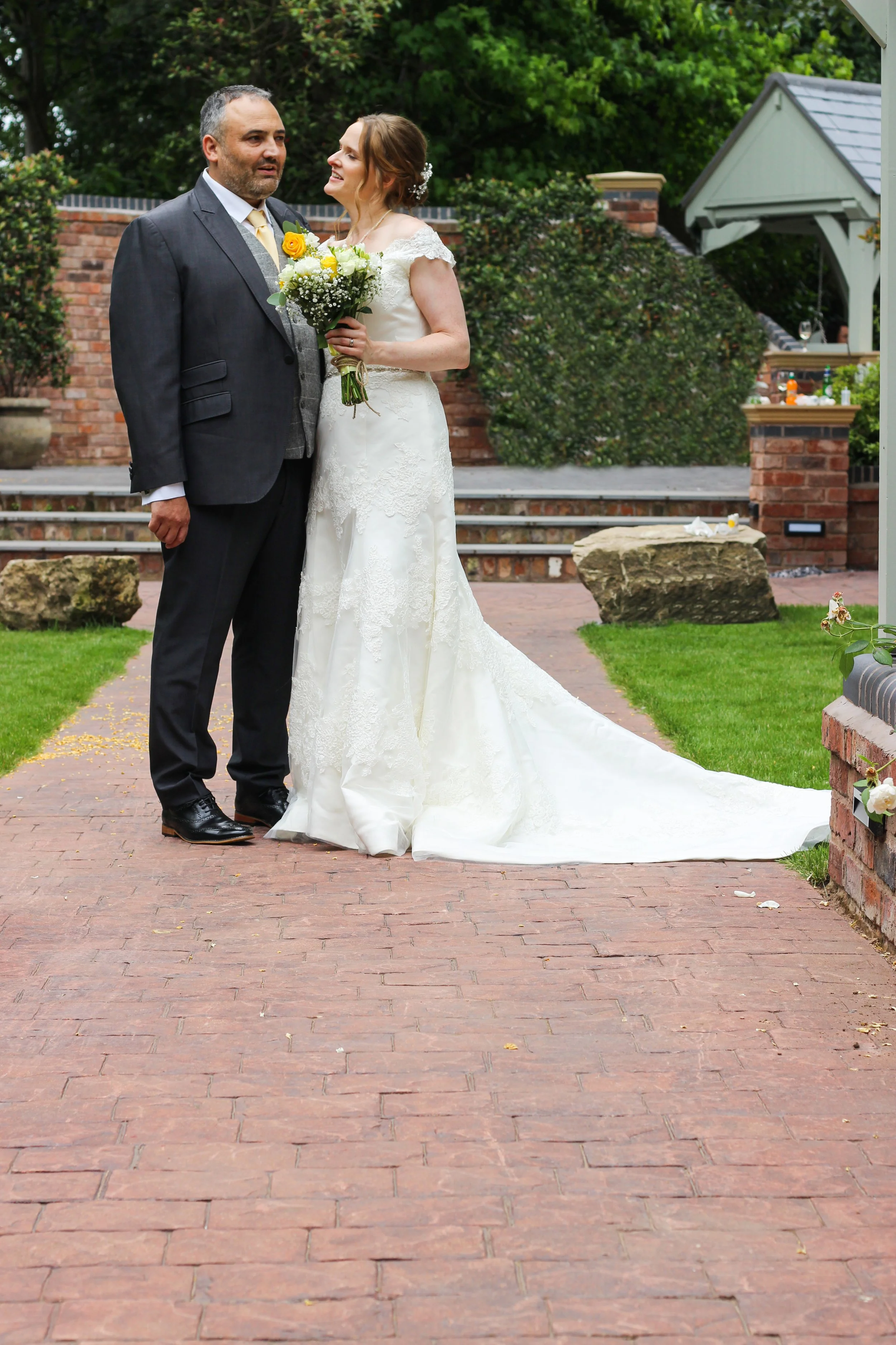 A bride and groom during their wedding, standing on a brick pathway outdoors. The bride is wearing a white lace wedding gown and holding a bouquet of yellow and white flowers. The groom is dressed in a dark suit with a light-colored vest and yellow t
