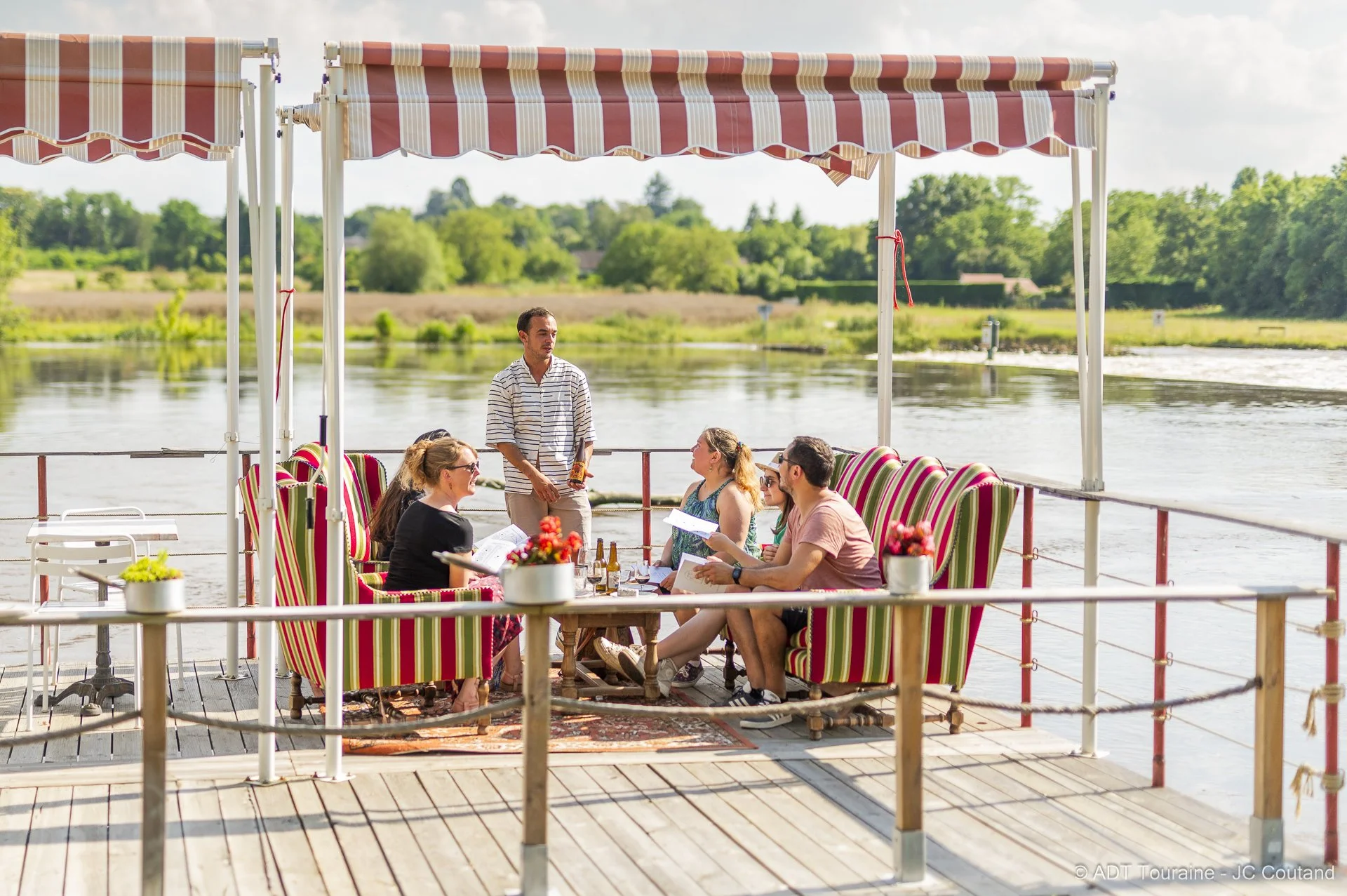 A group of people sitting and talking under a striped awning on a boat by a river, with greenery in the background.