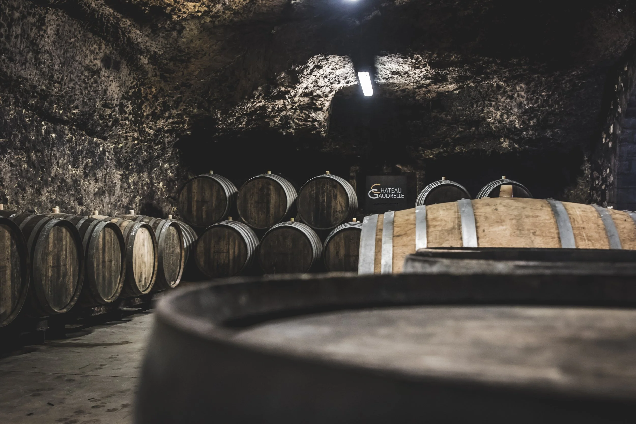 A wine cellar with rows of wooden barrels stored horizontally against a dark, rocky wall. There is a small black sign that reads 'Chateau Gaudrell' in the background.